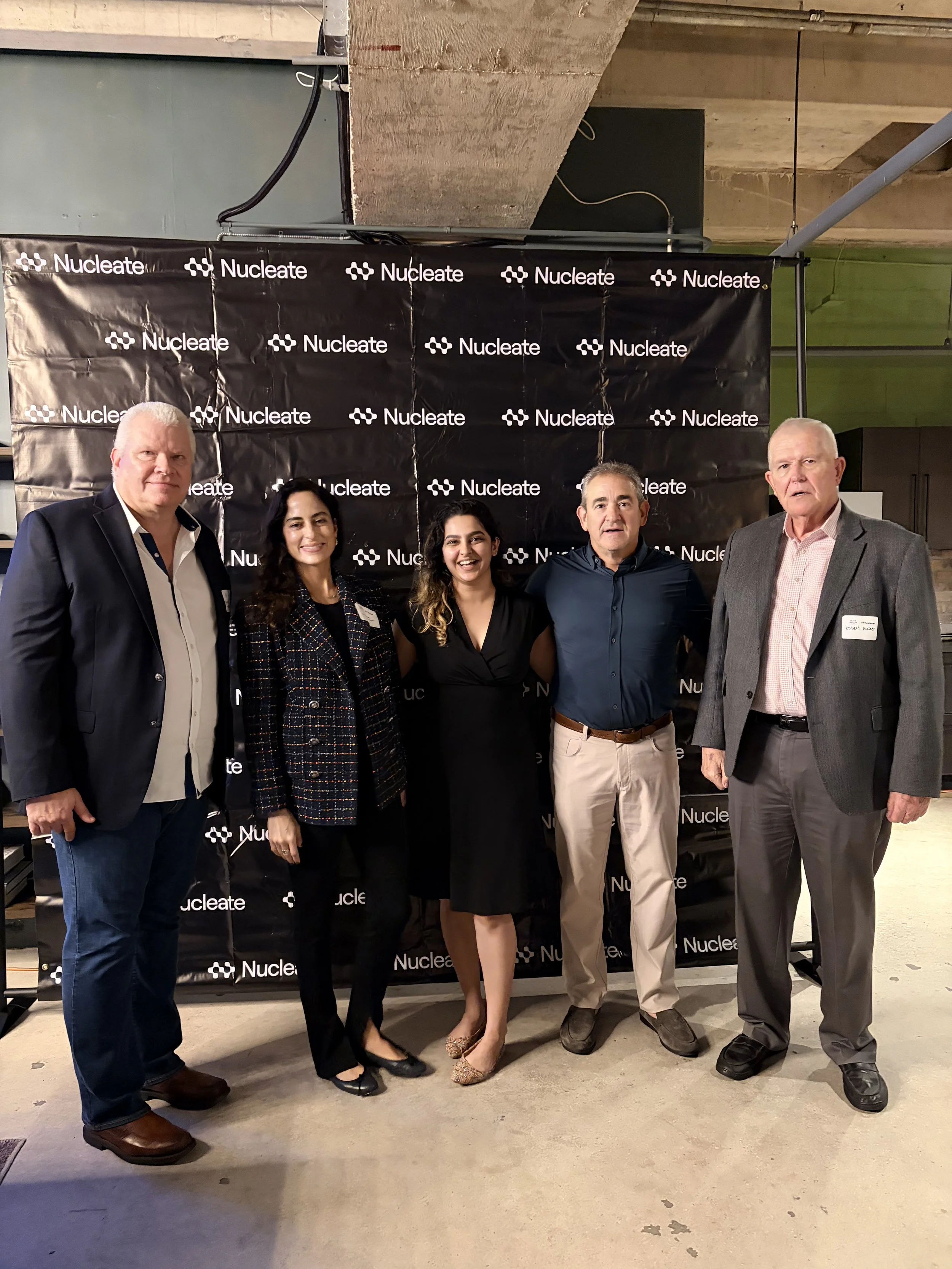 Five people standing in front of a black backdrop with the logo 'Nucleate.' The group includes three men and two women, dressed in business casual attire, smiling for the photo at an indoor event.