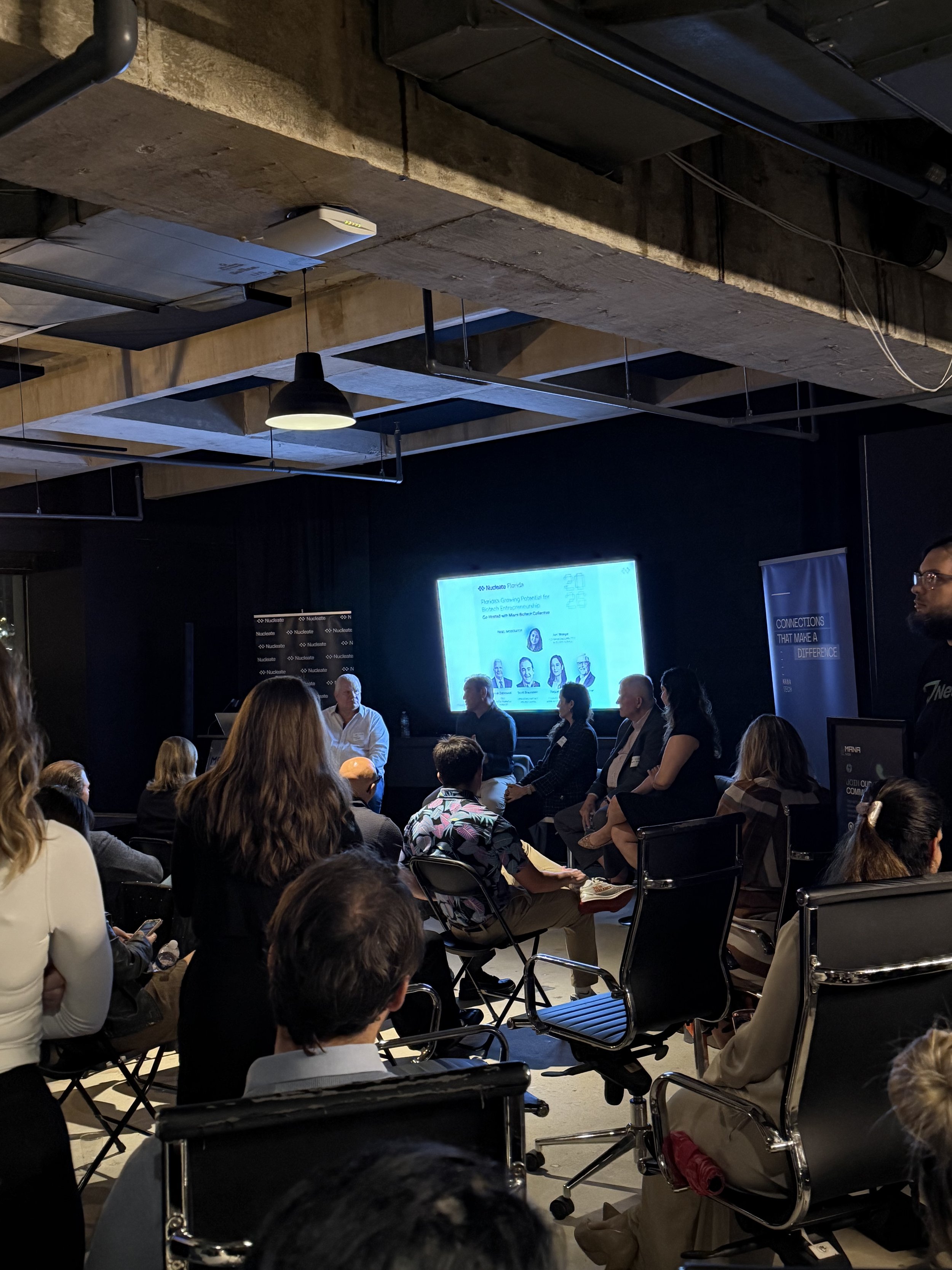 People attending a conference or presentation in a dark room with a large screen displaying a presentation slide.