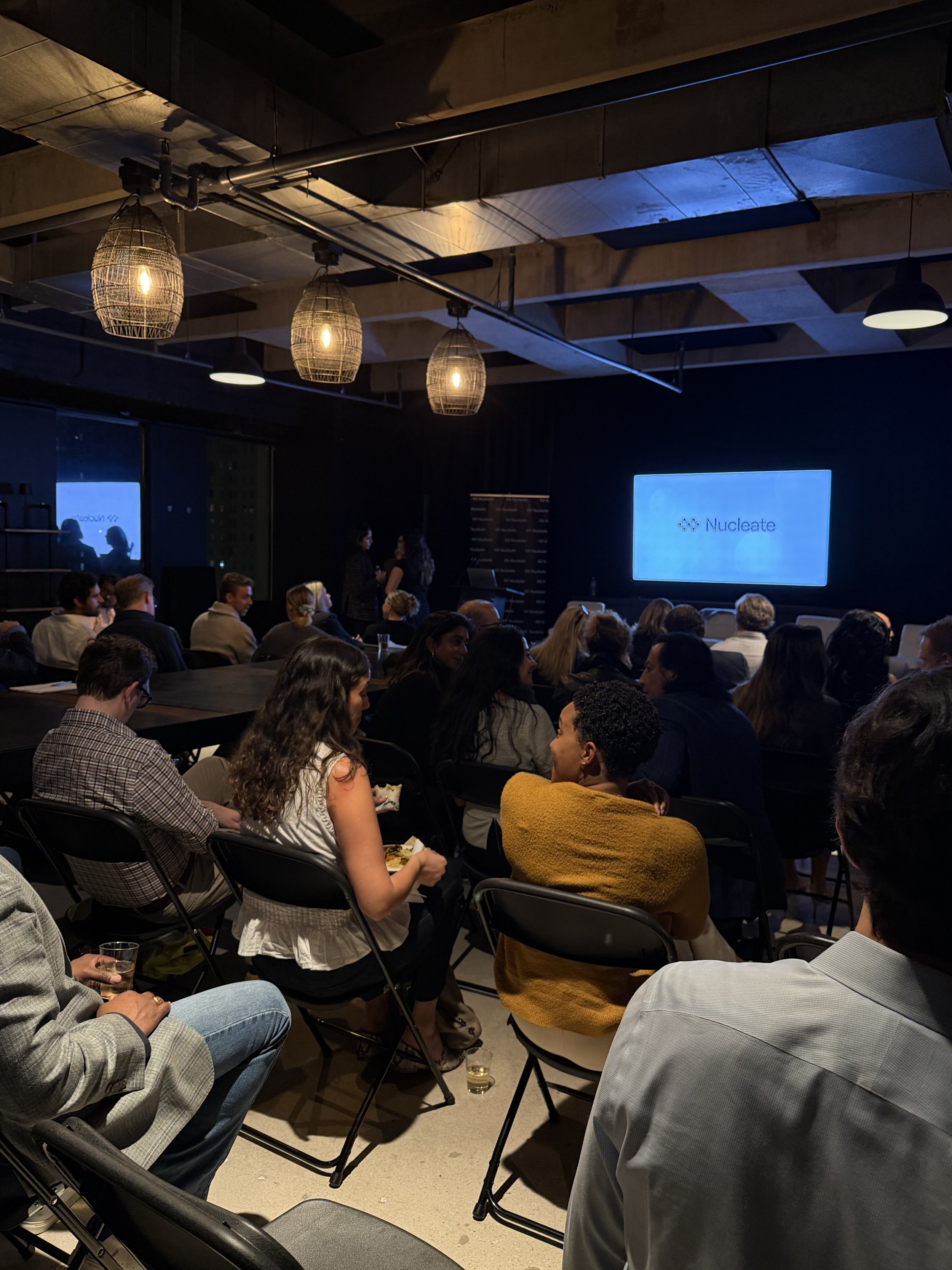 Indoor conference with people seated and standing, large screen displaying 'Nucleate' logo, ceiling lights, and an industrial ceiling design.