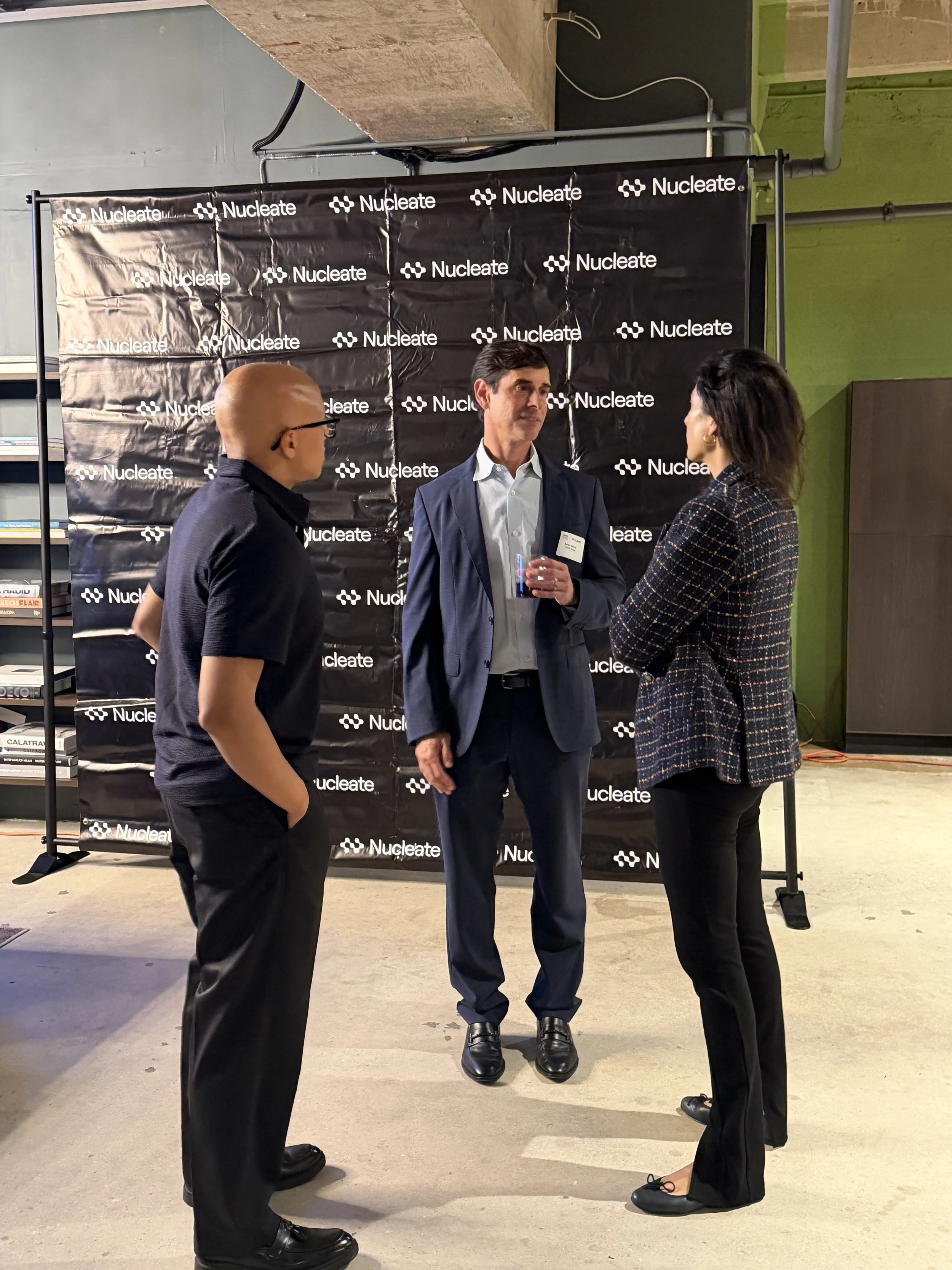 Three people are standing and talking in front of a black backdrop with the word 'Nucleate' repeated on it. The person in the middle is a man in a suit holding a drink, and two women are on either side of him, engaged in conversation.