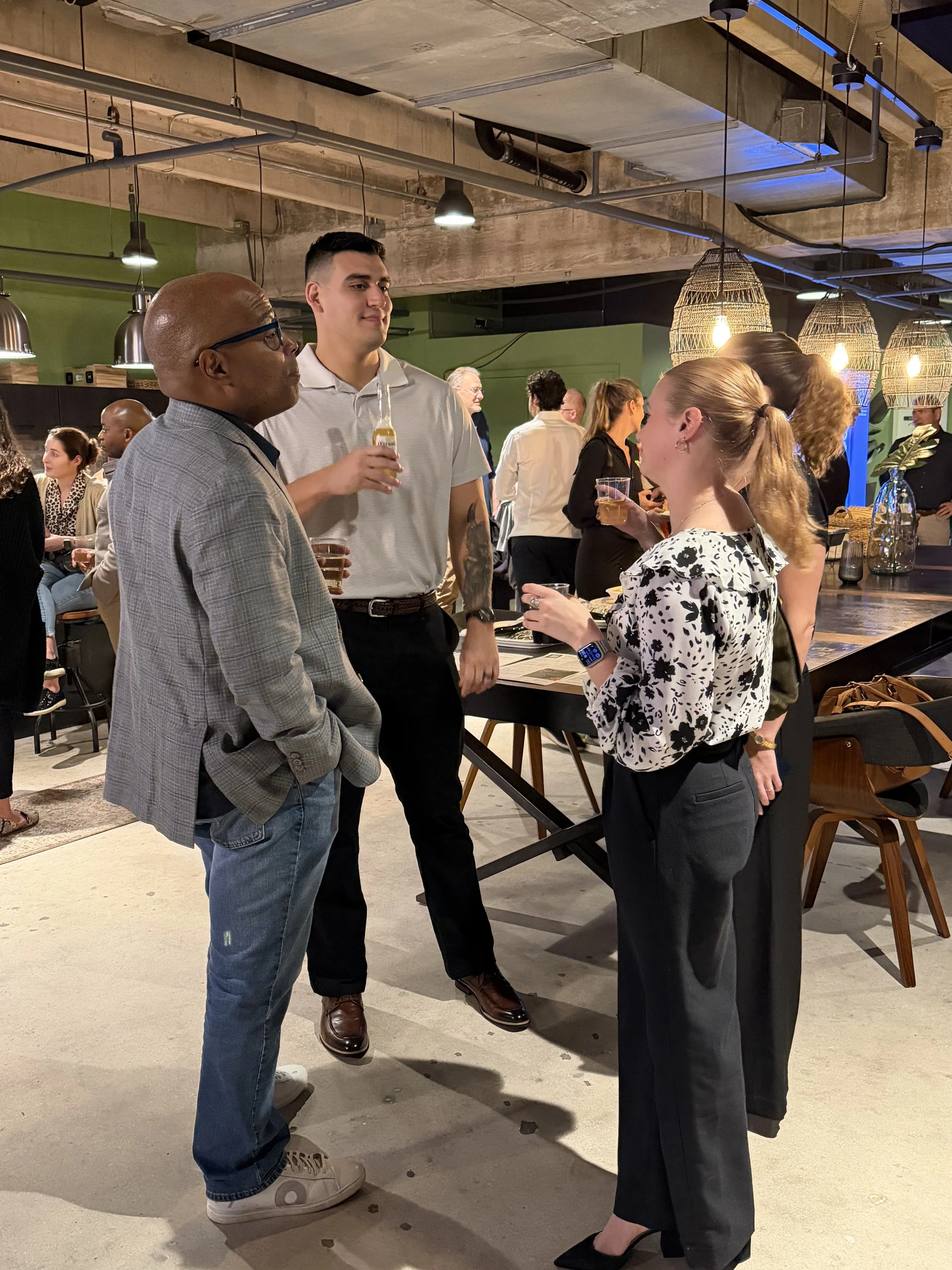 People socializing at a bar or restaurant, engaging in conversation, with some holding drinks, in an industrial-style interior with hanging wicker lights.