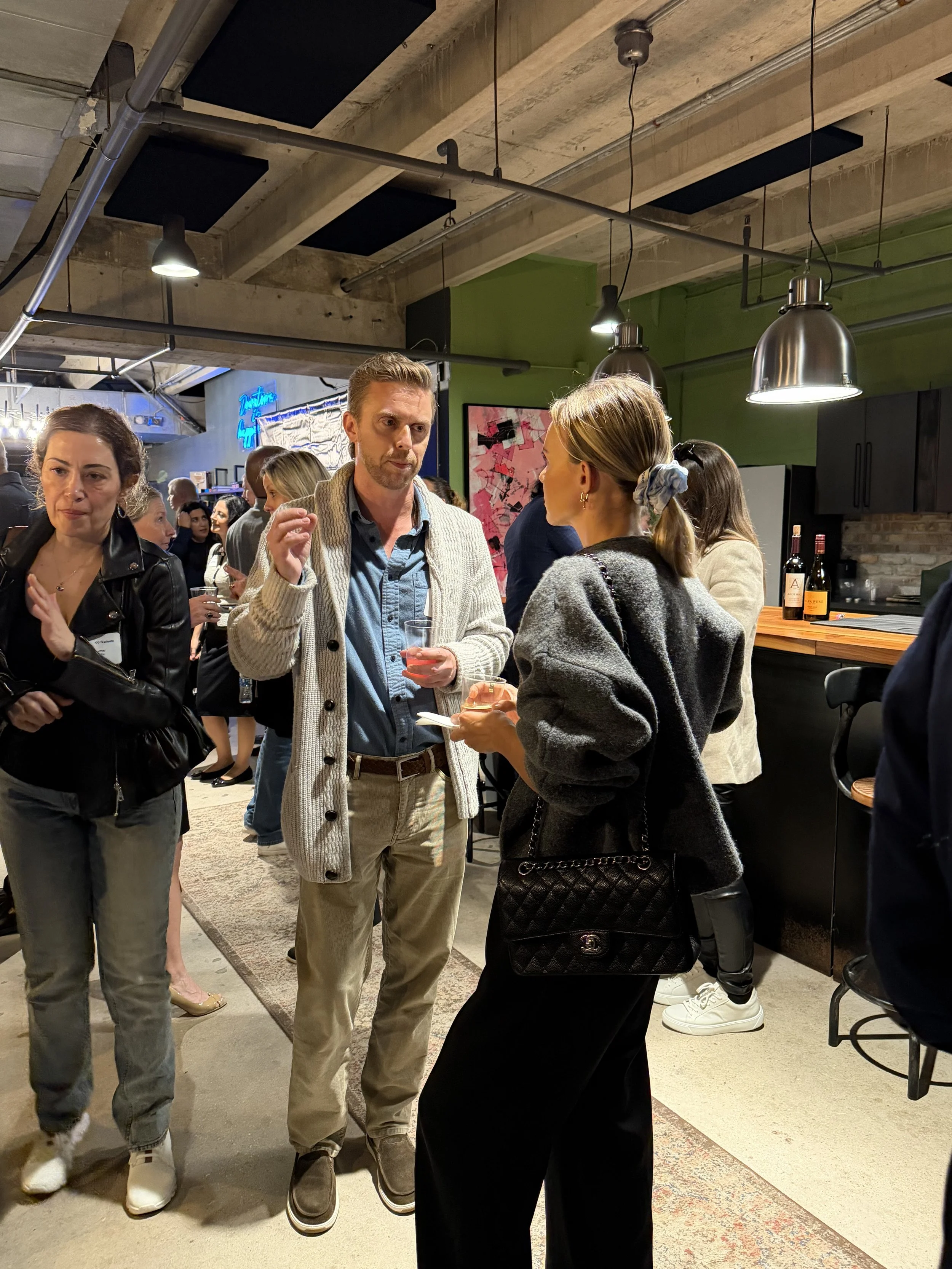 Group of people socializing in a modern, industrial-style bar or restaurant with concrete ceiling, black pendant lights, and green wall decor.