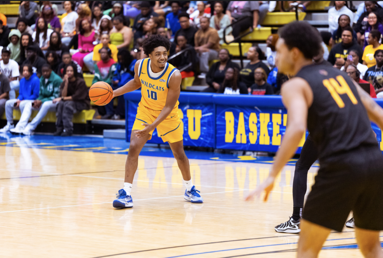 A basketball player in a yellow uniform with the number 10 dribbling, facing an opponent in black jersey with number 14, on a basketball court with an audience in the background.