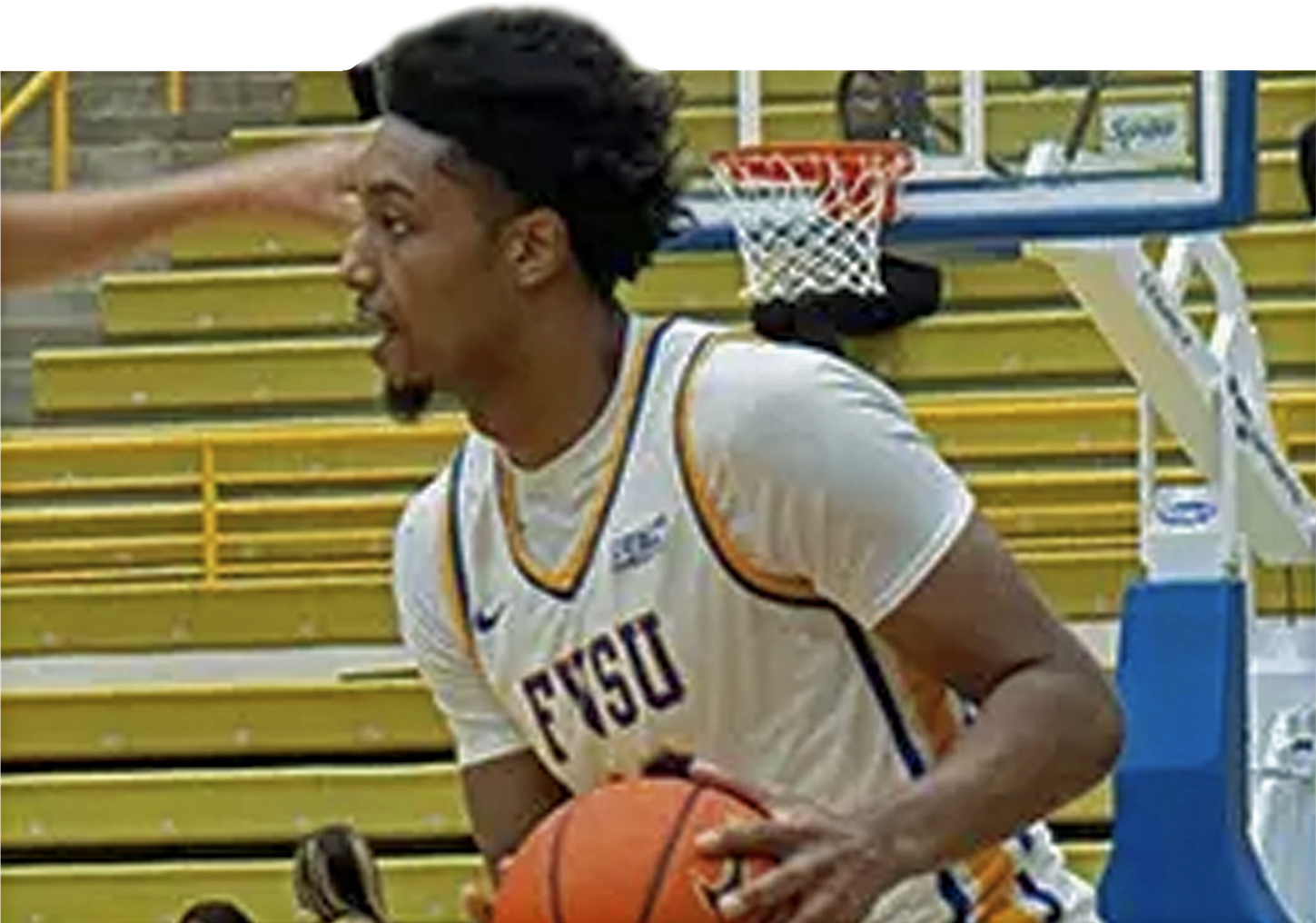 A basketball player in a Fresno State uniform holding a basketball during a game or practice, with yellow bleachers and a basketball hoop in the background.
