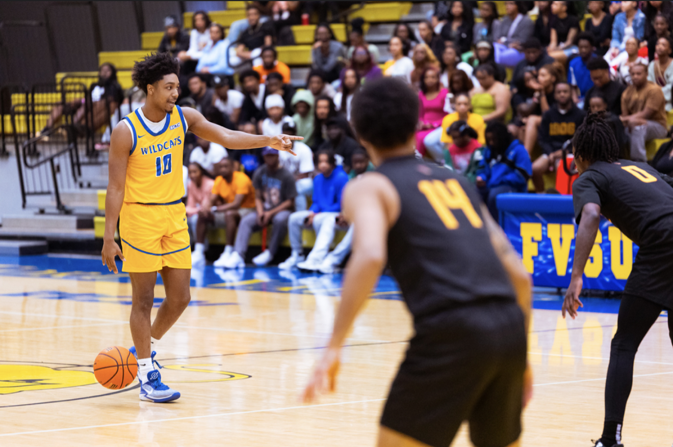 A women's basketball game with players on the court and an audience in the background. One player in a yellow uniform with number 10 has the basketball, while two players in black uniforms with numbers 14 and 0 are defending.