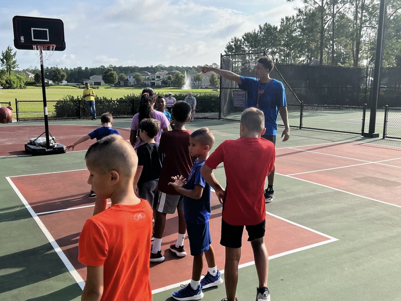 Young children lining up on an outdoor basketball court while a coach in a blue shirt instructs them. Basketball hoops and a black fence surround the court, with trees and houses in the background.
