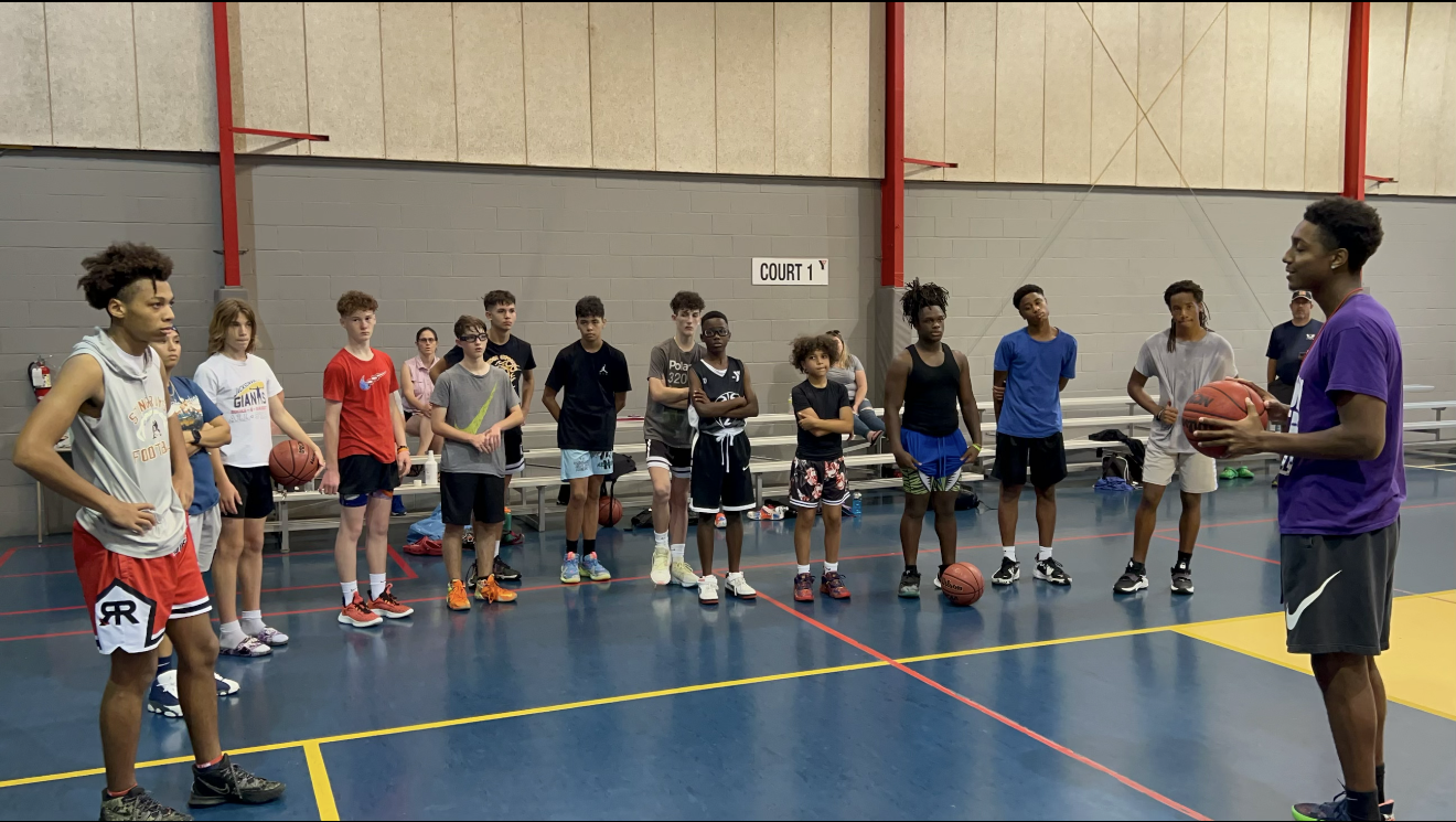 Basketball coach holding a ball speaking to a group of young players standing in a row in an indoor gymnasium.