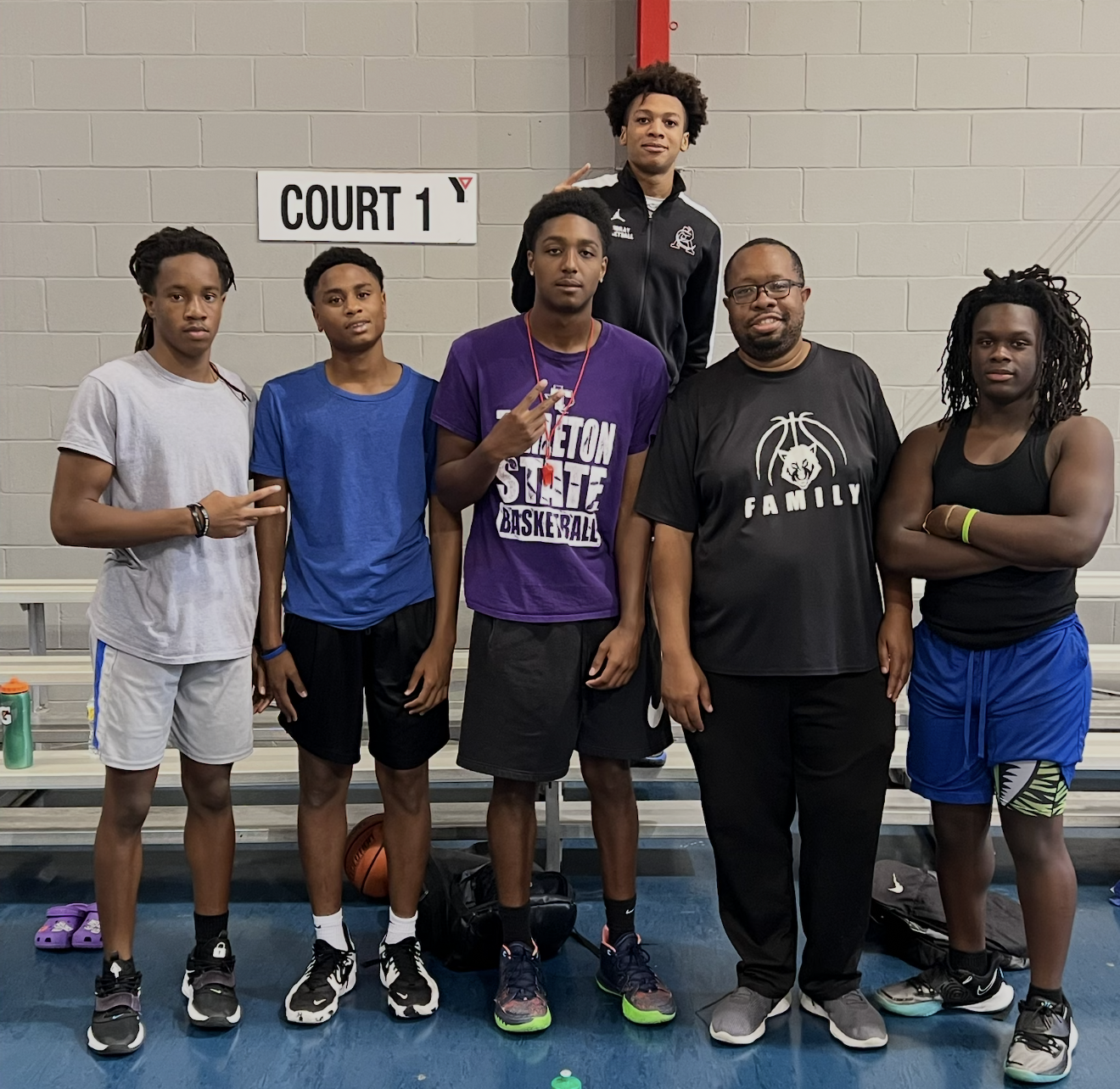 Group of six young men and one adult male standing in front of a sign that says 'Court 1' in a gymnasium. The group is posed for a photo, some making hand gestures, with sports bags and water bottles on the bench behind them.