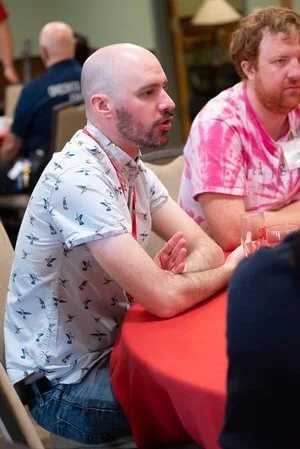 A bald man with a beard wearing a white shirt with a blue pattern sitting at a round table with a red tablecloth, in a social setting with other people in the background.