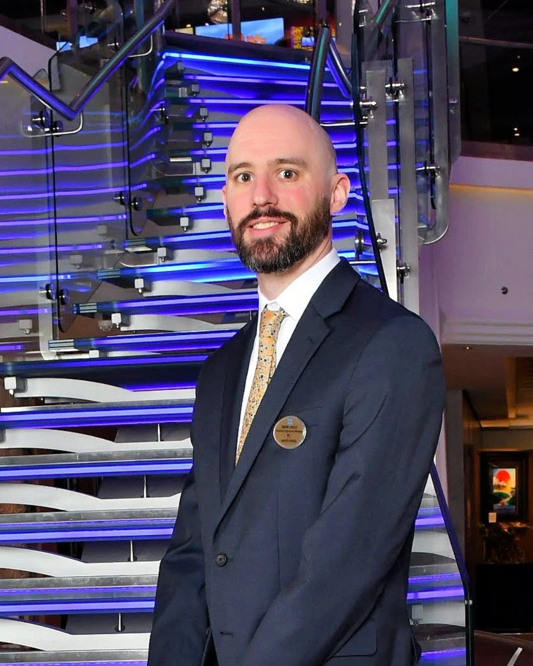 A man in a dark suit with a badge poses in front of a brightly lit, modern staircase with blue LED lights.