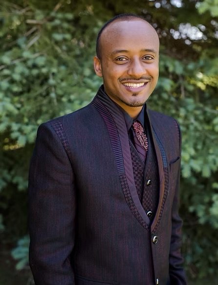 A smiling man in a dark suit standing outdoors with green foliage in the background.