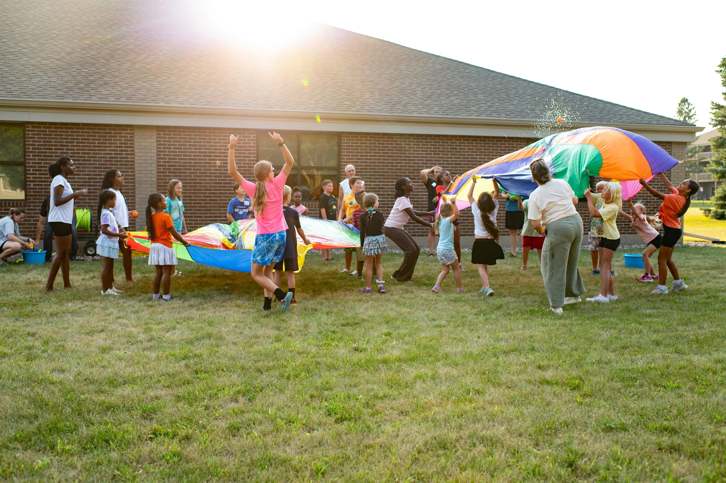 Children and adults playing with a large colorful parachute outdoors in front of a brick building. The sun is shining brightly in the sky.