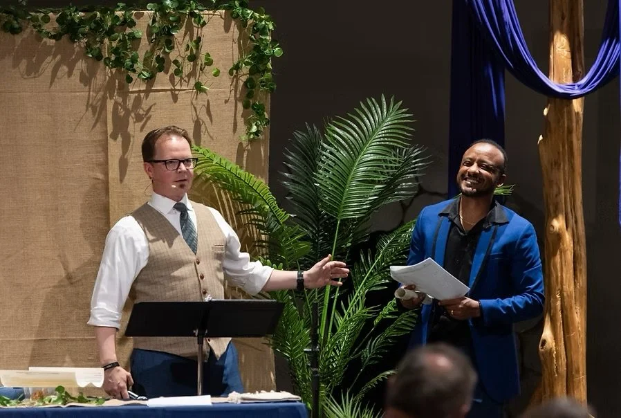 Two men stand on a stage near a large green plant, one holding a microphone and paper, the other with glasses and a vest, engaged in conversation, with a backdrop of beige and dark walls and a purple curtain.