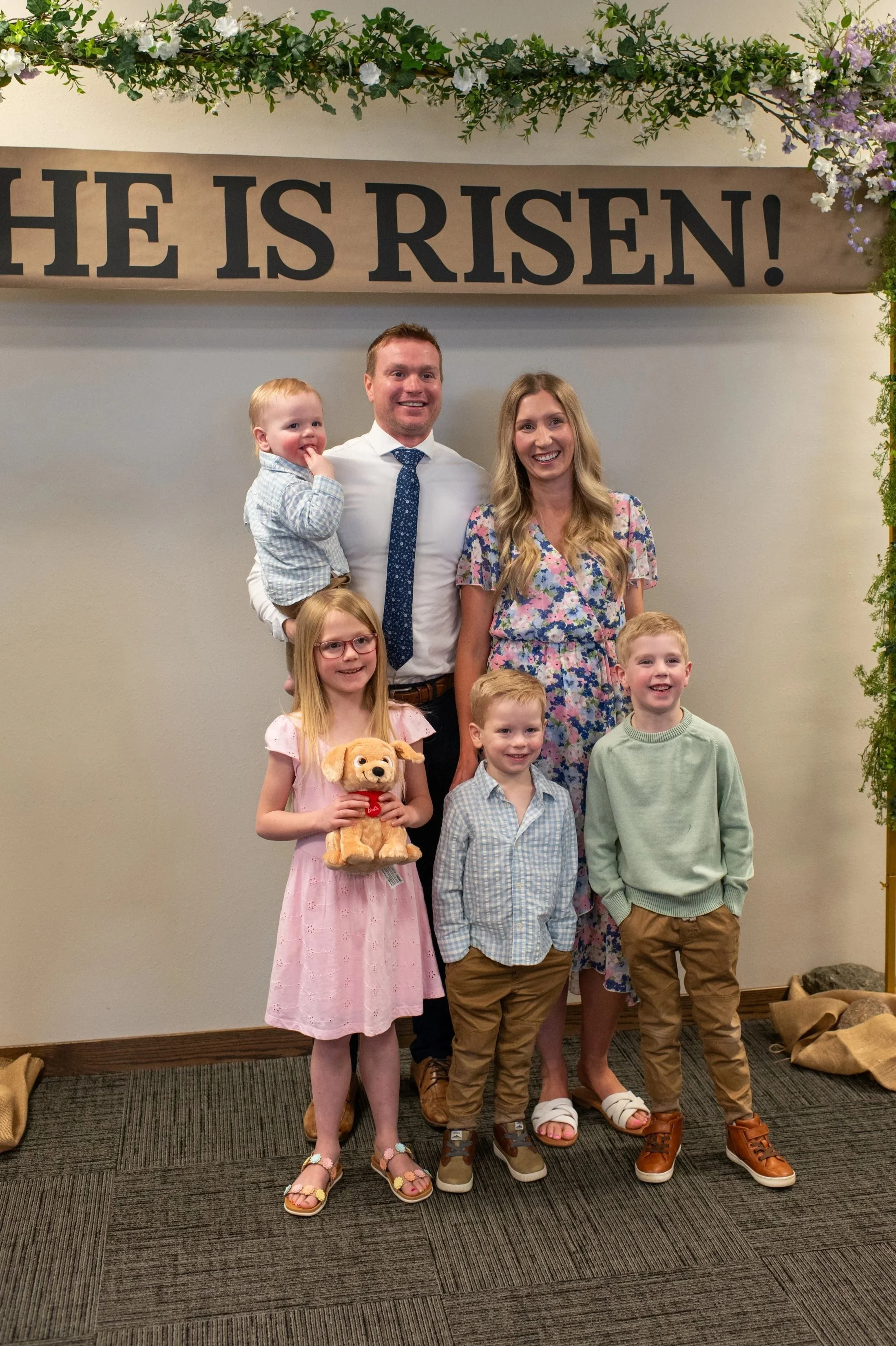 People standing in front of a sign that says 'He is Risen!' with flower decorations above. The group includes a woman, a man, and four children, some smiling and one holding a stuffed animal.