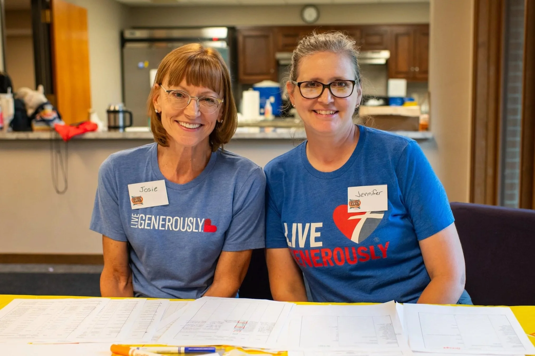 Two women sitting at a table in a kitchen, wearing blue T-shirts with the words 'Live Generously' and name tags, smiling at the camera.