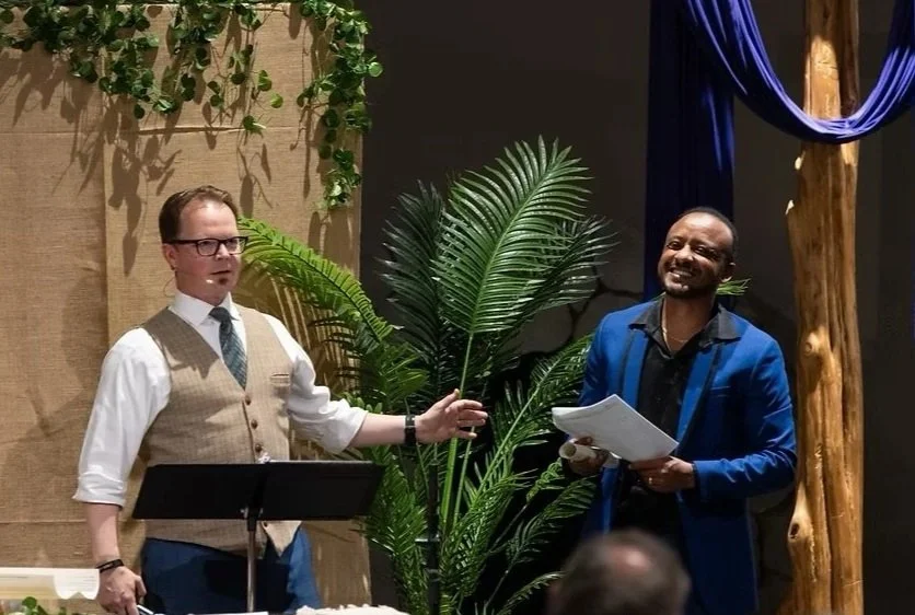 Two men are standing in front of a large plant; one is holding a palm leaf and the other is smiling while holding papers. They appear to be in a formal or staged setting, possibly during a presentation or performance.