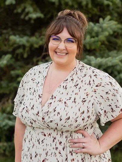 A woman with glasses and brown hair styled in a bun, smiling outdoors with greenery in the background.
