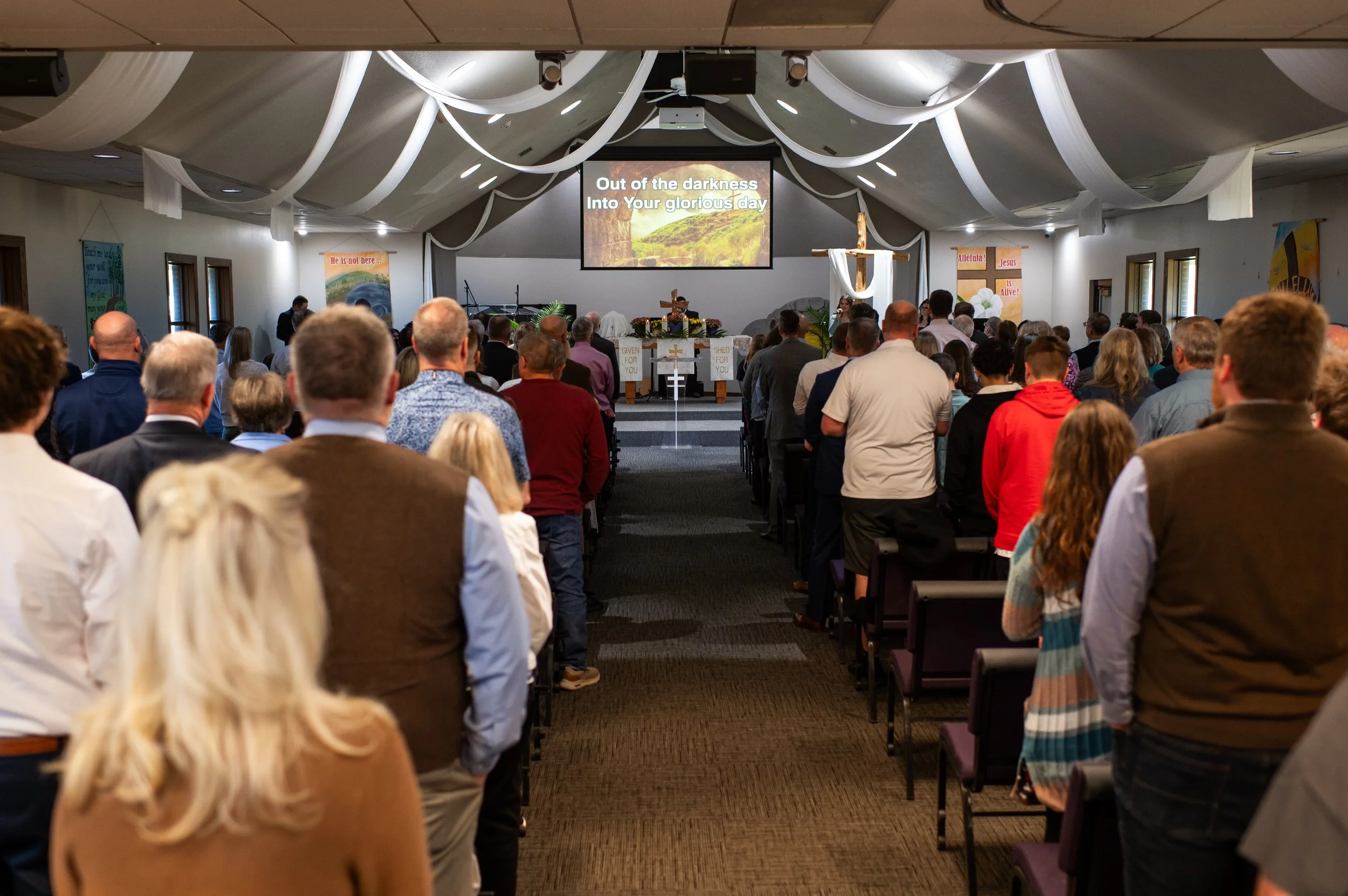A congregation inside a church during a service, facing an altar with a cross, floral decorations, and a large screen displaying lyrics, with white fabric drapes hanging from the ceiling.