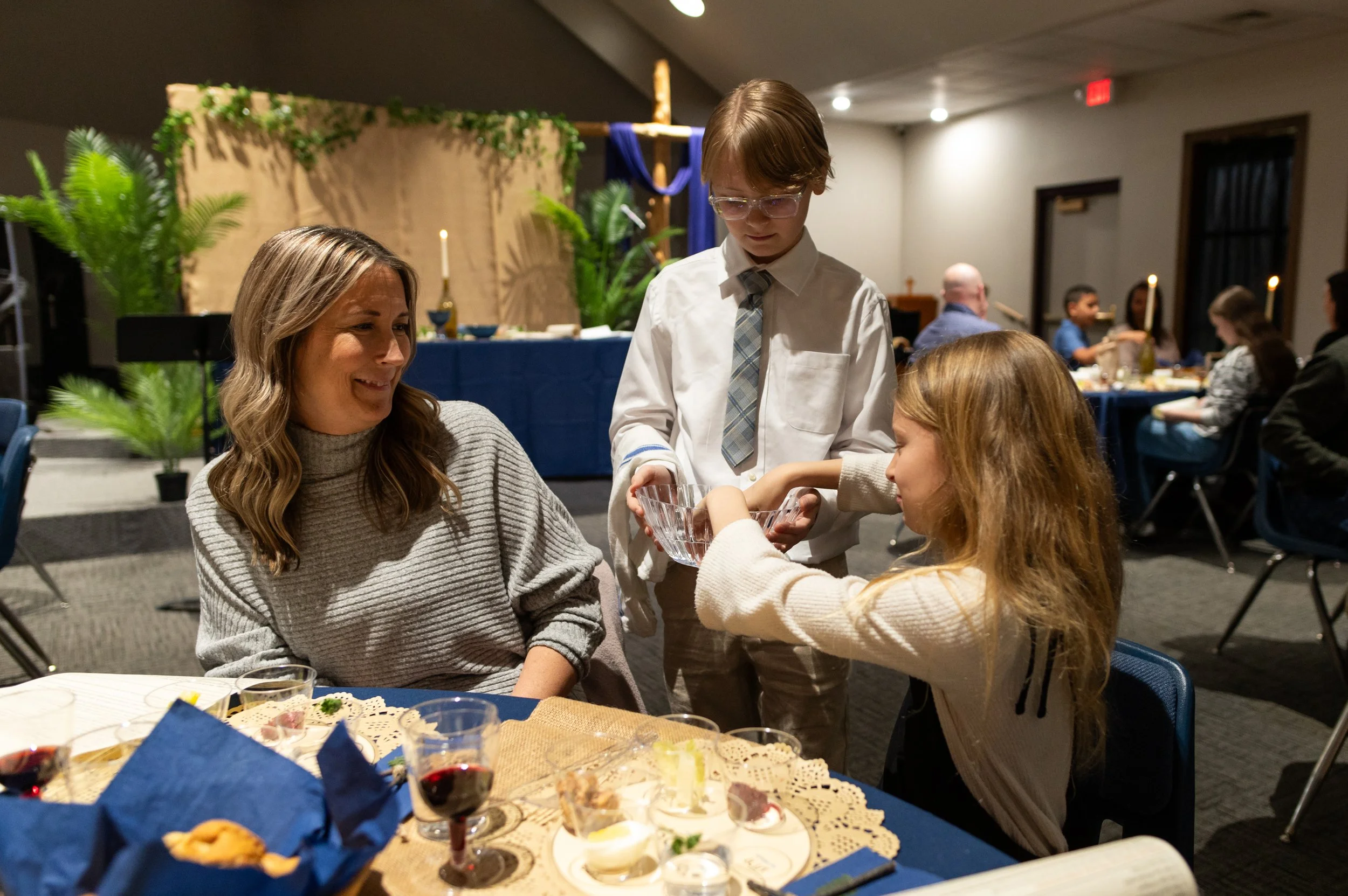 A group of children and adults at a festive gathering, with food and drinks on the table. One child is serving a glass of water to a woman seated at the table. The background includes a decorated setup with candles and greenery.