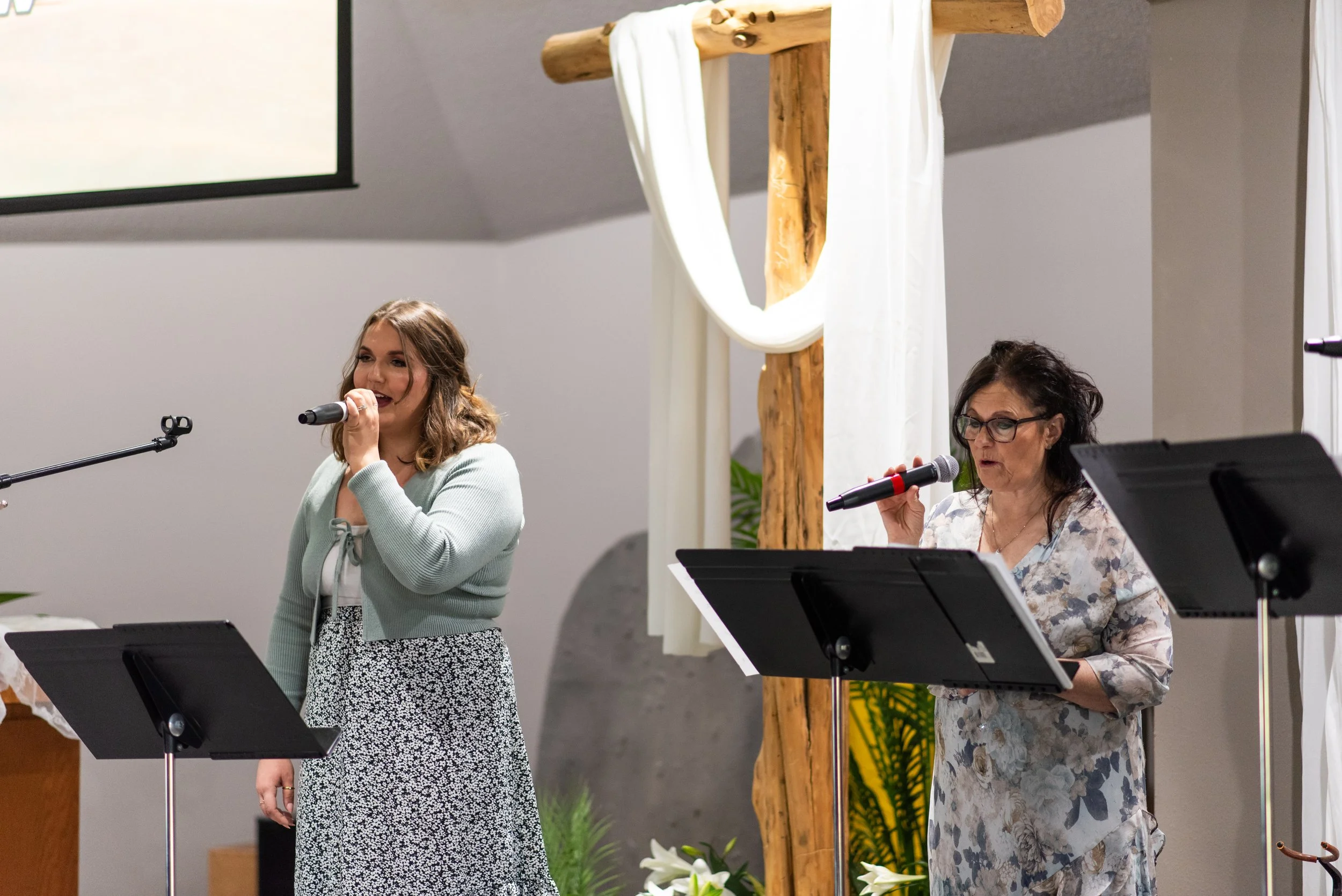 Two women are speaking or singing into microphones at a church or event setting, with music stands in front of them and a decorative wooden cross behind them.