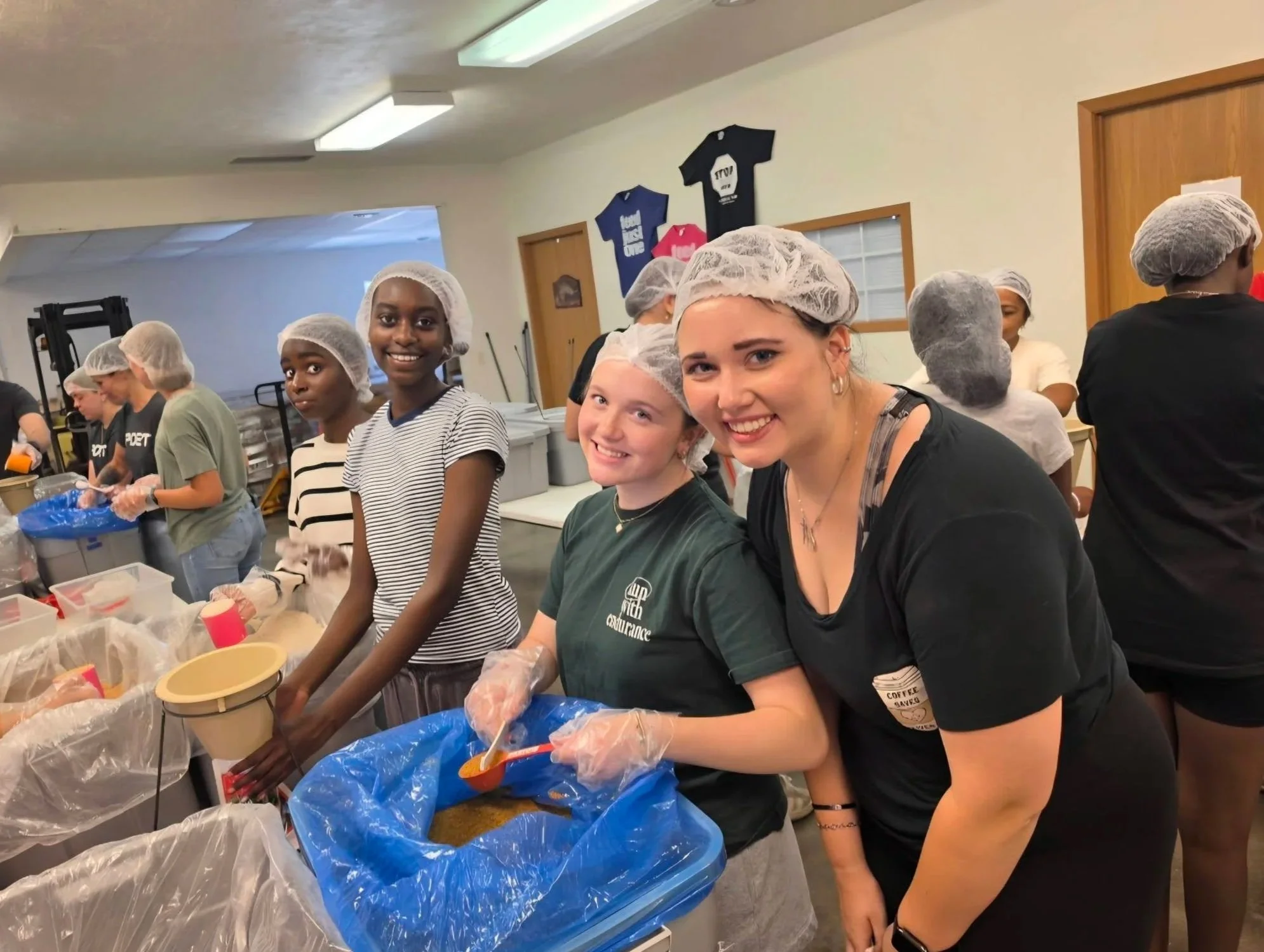 A group of diverse young women and girls wearing hairnets and gloves, working together in a kitchen or food preparation area, preparing food or ingredients on a table covered with plastic bags and containers.