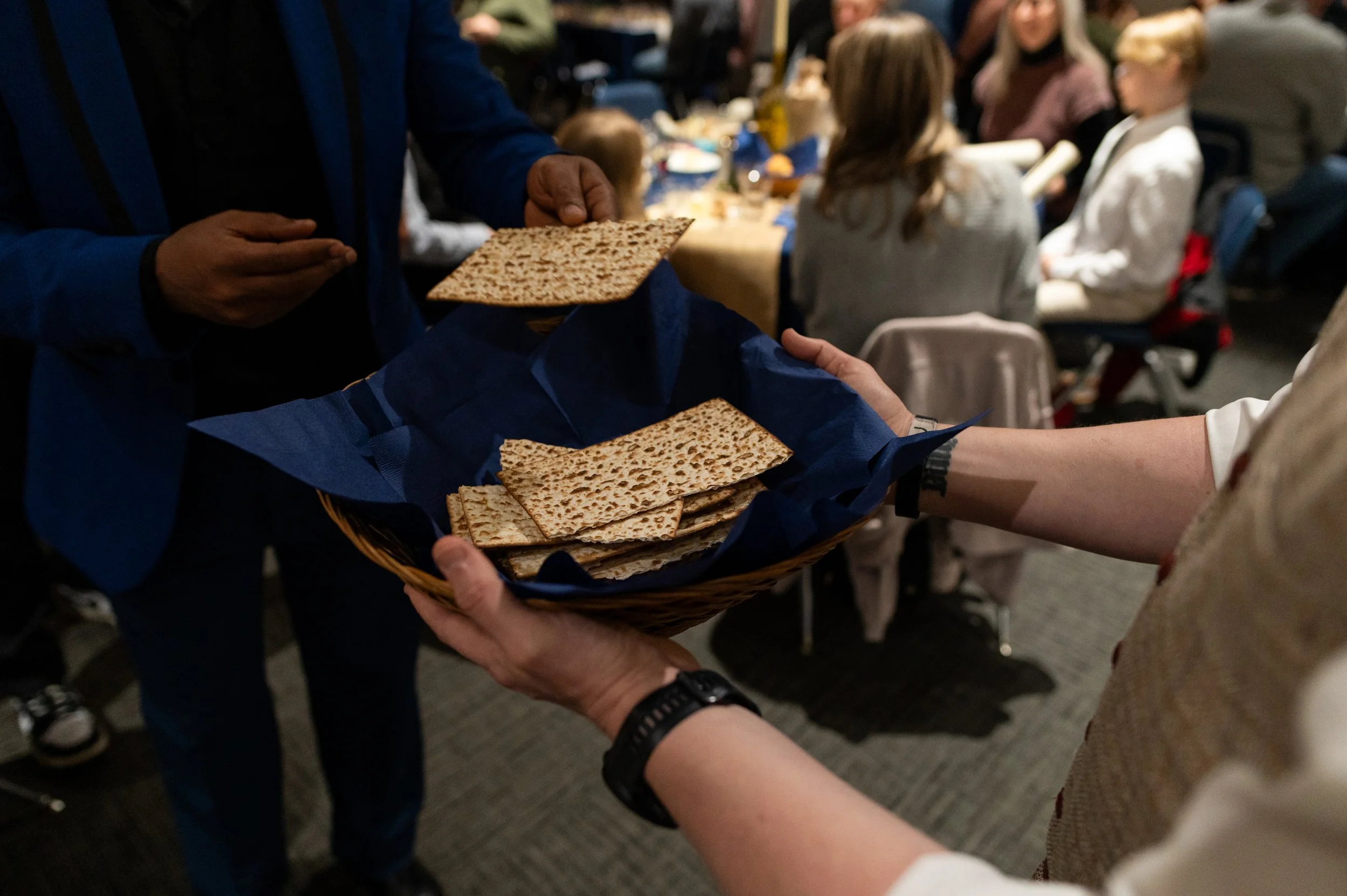 Person in a blue jacket is handing a basket of matzah to another person in a beige shirt during a gathering.