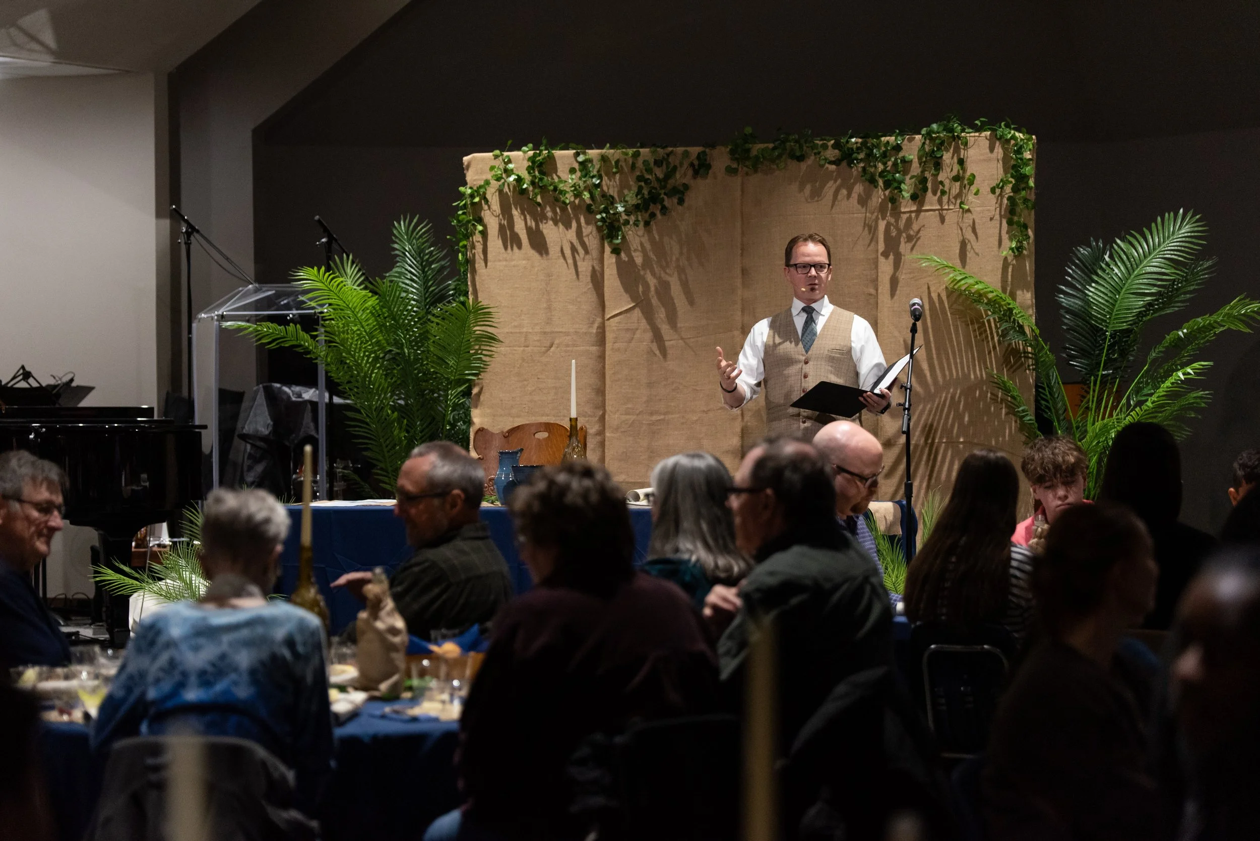A man in a vest and tie speaking at a microphone during an indoor event, with green plants and a beige backdrop behind him, and a crowd seated in front.