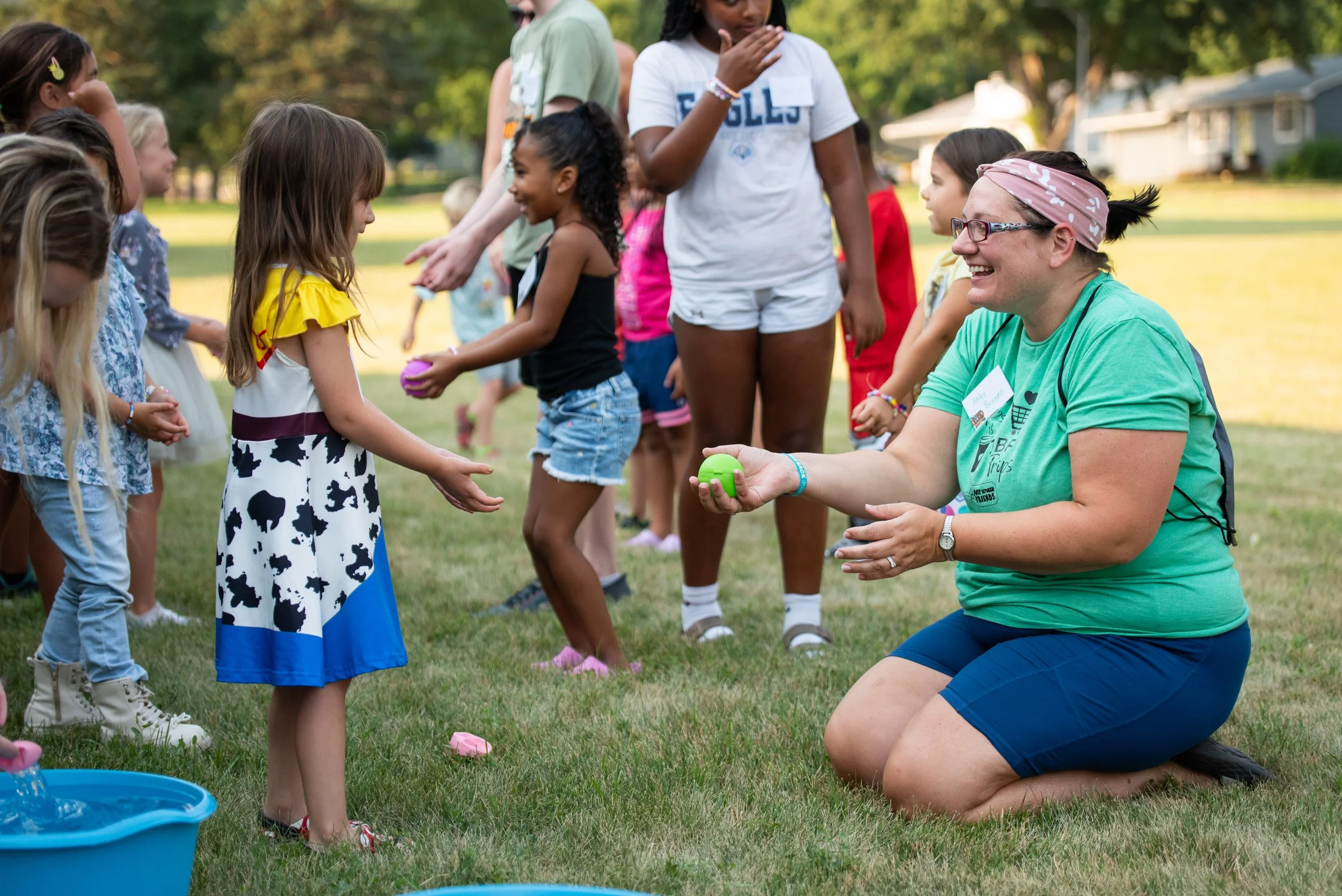 A group of children and an adult woman outdoors on grass. The woman is squatting and smiling, holding a green egg, while children stand in a line, participating in an activity. Trees and a house are in the background.