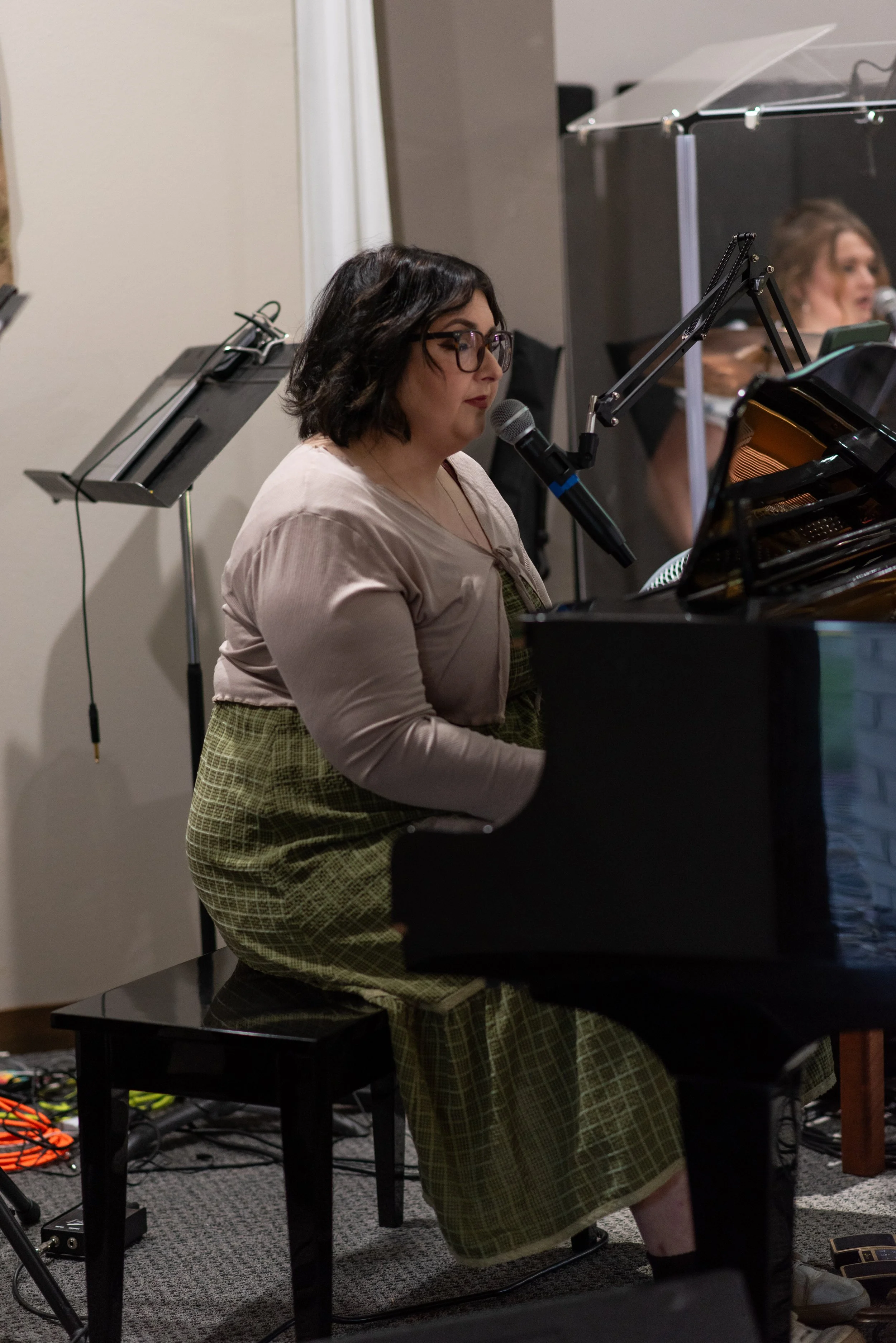 A woman with short dark hair, glasses, and a light-colored top, playing piano at a music performance or rehearsal. There is another person in the background singing into a microphone.