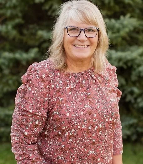 Smiling woman with blonde hair, glasses, wearing a pink floral long-sleeve top, standing outdoors with greenery in the background.