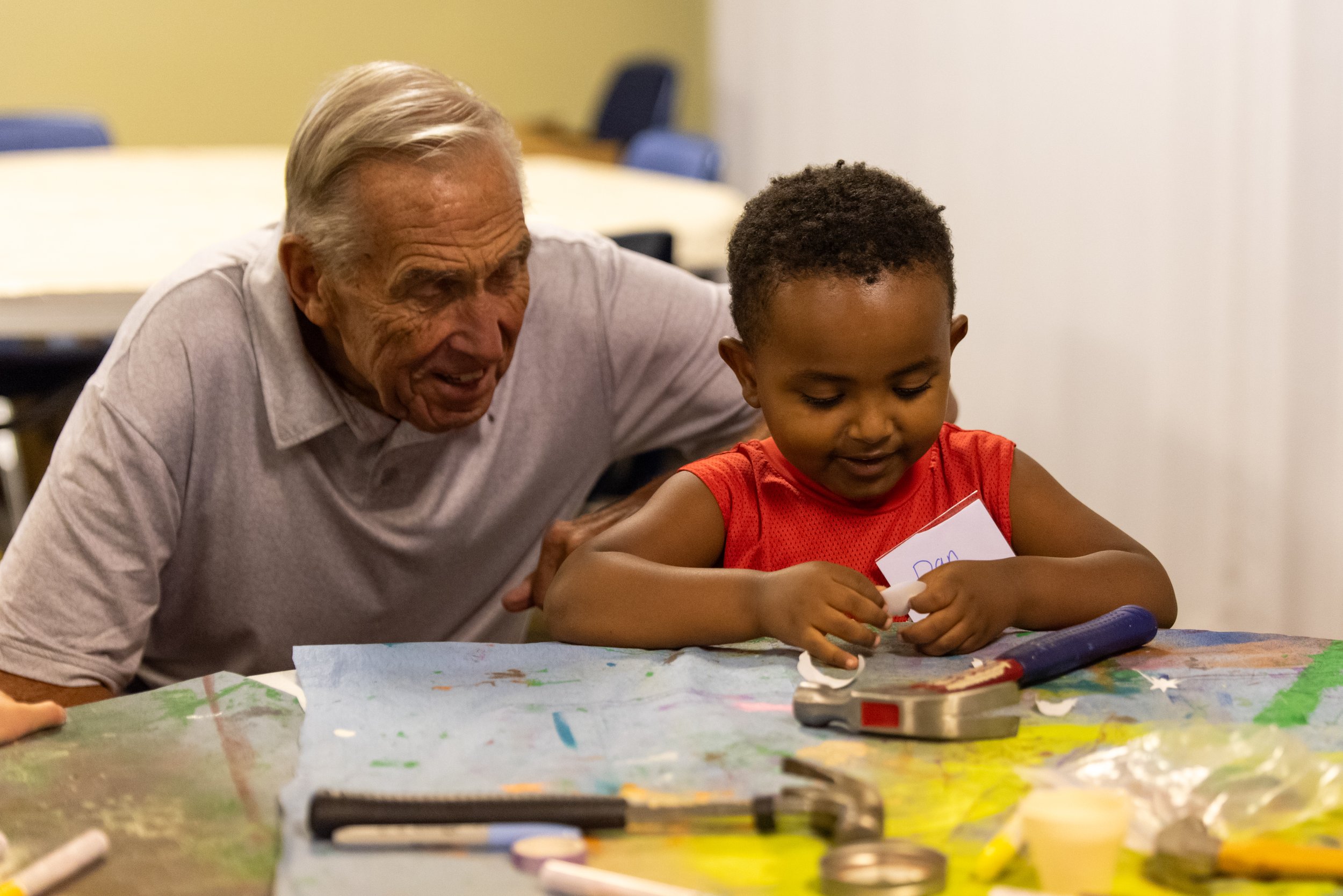 An elderly man and a young boy sitting at a colorful table. The elderly man is leaning in and smiling, while the boy is focused on drawing or writing on a piece of paper. The table has art supplies and colorful drawings.