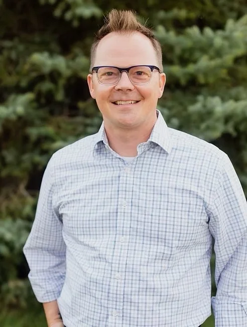A smiling man with glasses wearing a checkered button-up shirt outdoors with green trees in the background.