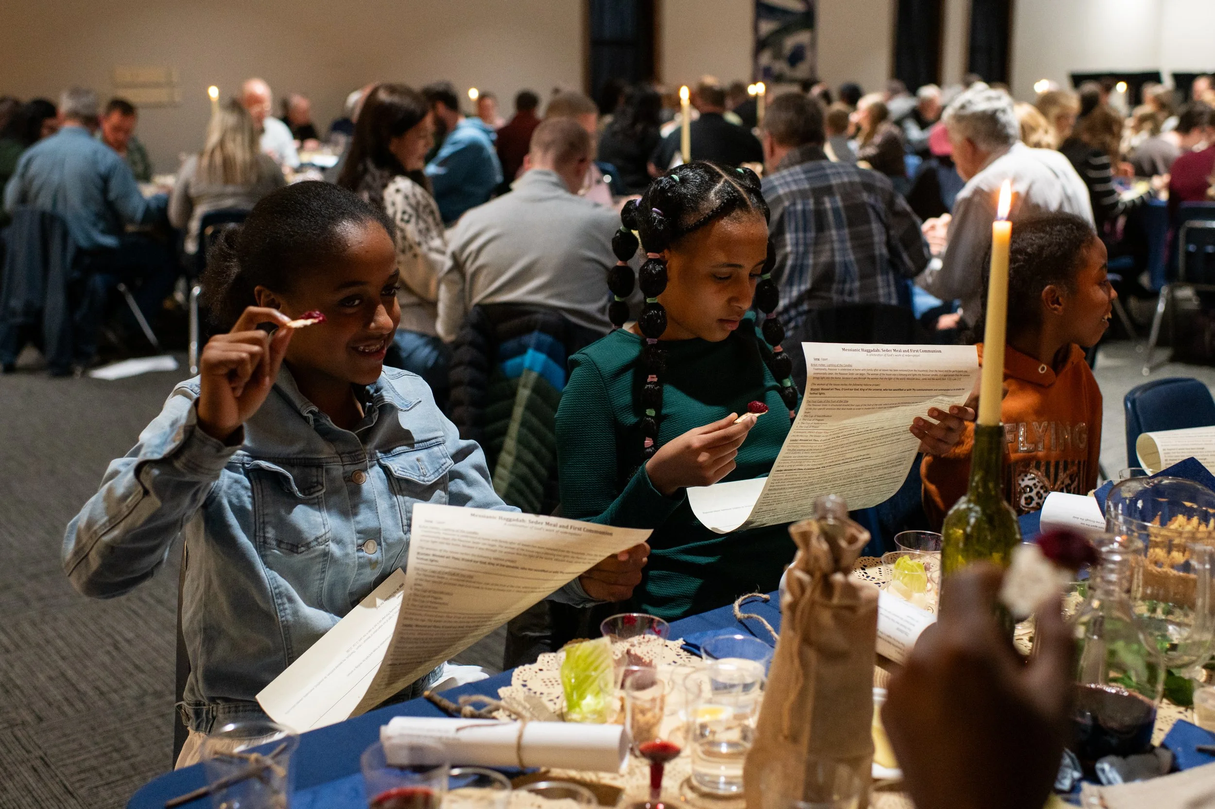 A group of people, including children and adults, sitting at a long dining table with food and candles in a dimly lit room, likely at a celebration or gathering.