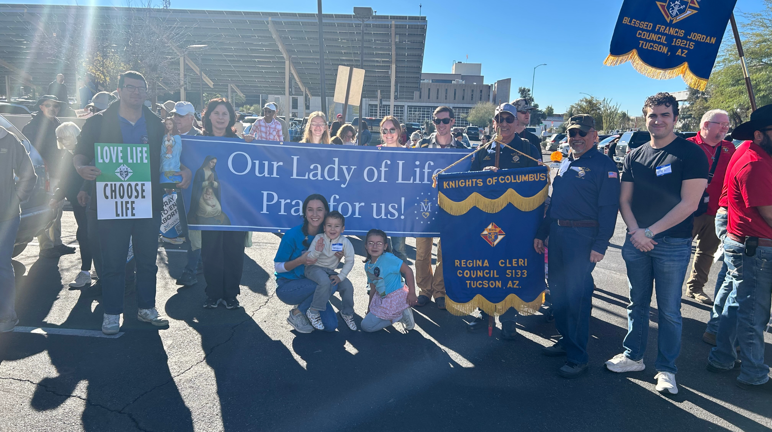 Group of people at a parade holding banners for Our Lady of Life and Knights of Columbus, Tucson, AZ. Some are smiling, with children in front, and a blue sky overhead.