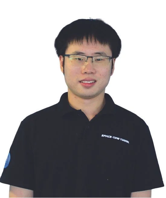 Professional portrait of a young man with short black hair, wearing glasses, a gray checkered blazer, and a black shirt, standing against a plain light-colored background.