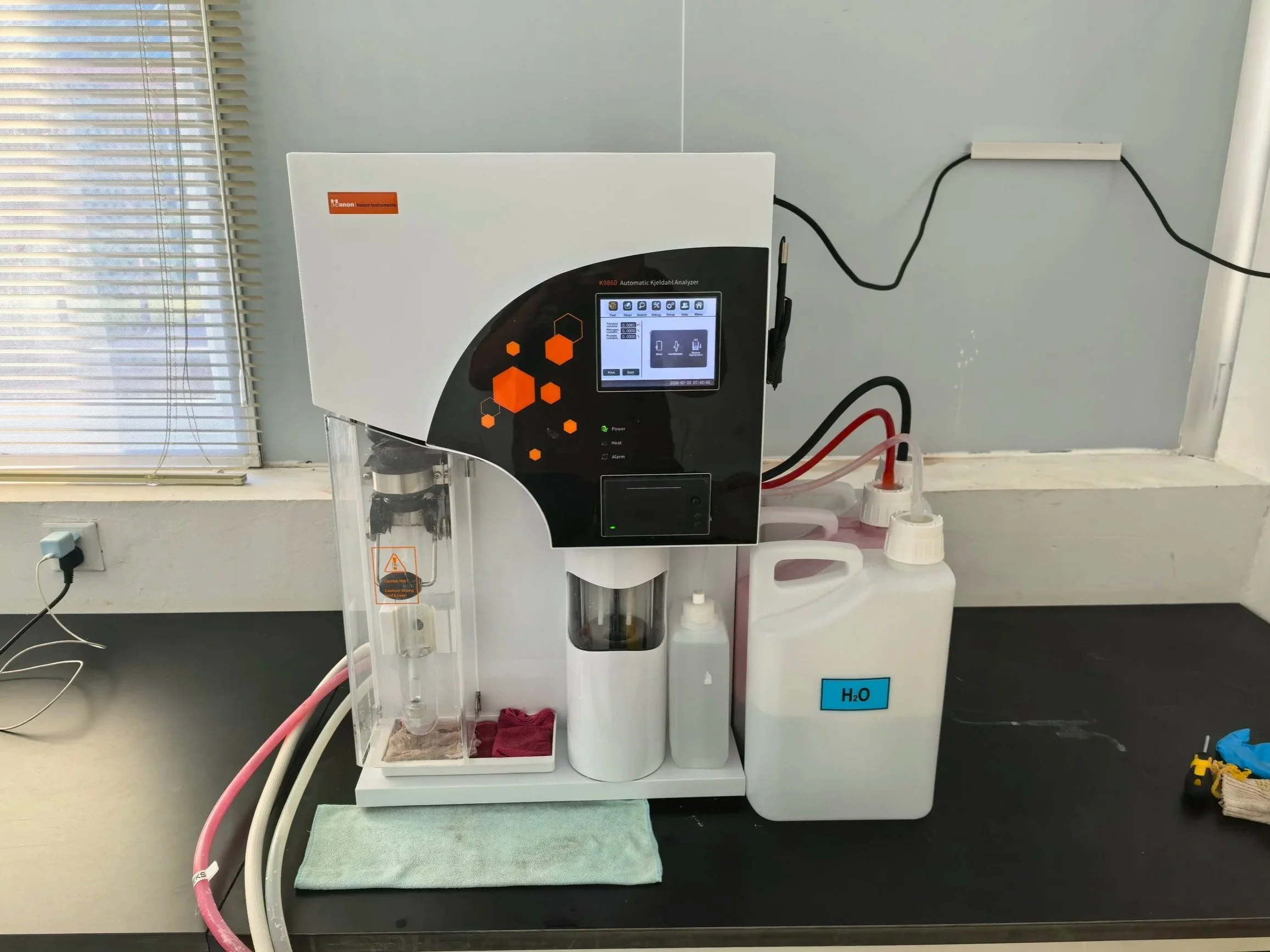 A laboratory setup with a white machine labeled 'Kanon K9860 Automatic Kjeldahl Analyzer' on a countertop, electrical cords, and various bottles including a large container labeled 'H2O'. There are blinds covering a window in the background.