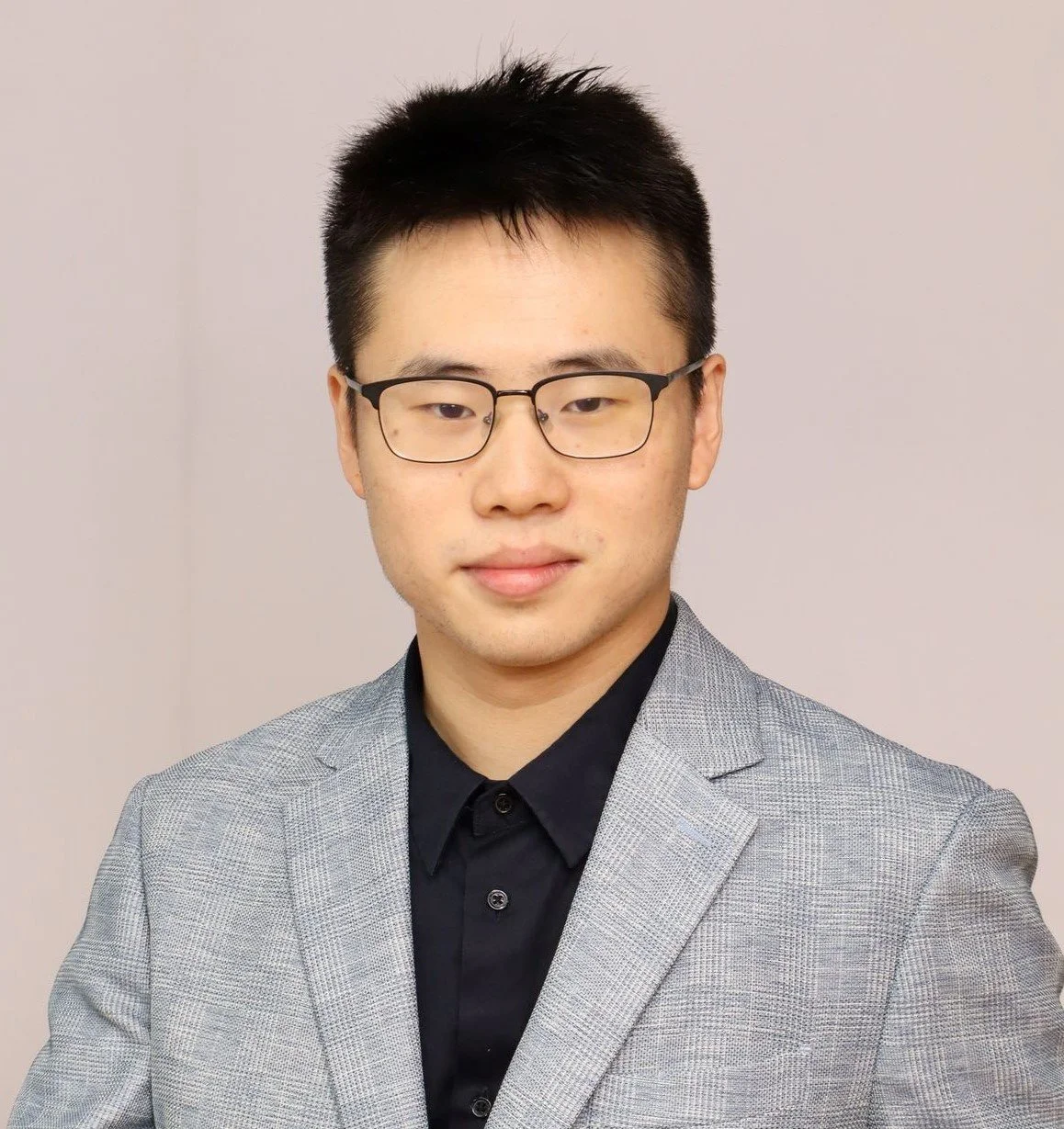 Professional portrait of a young man with short black hair, wearing glasses, a gray checkered blazer, and a black shirt, standing against a plain light-colored background.