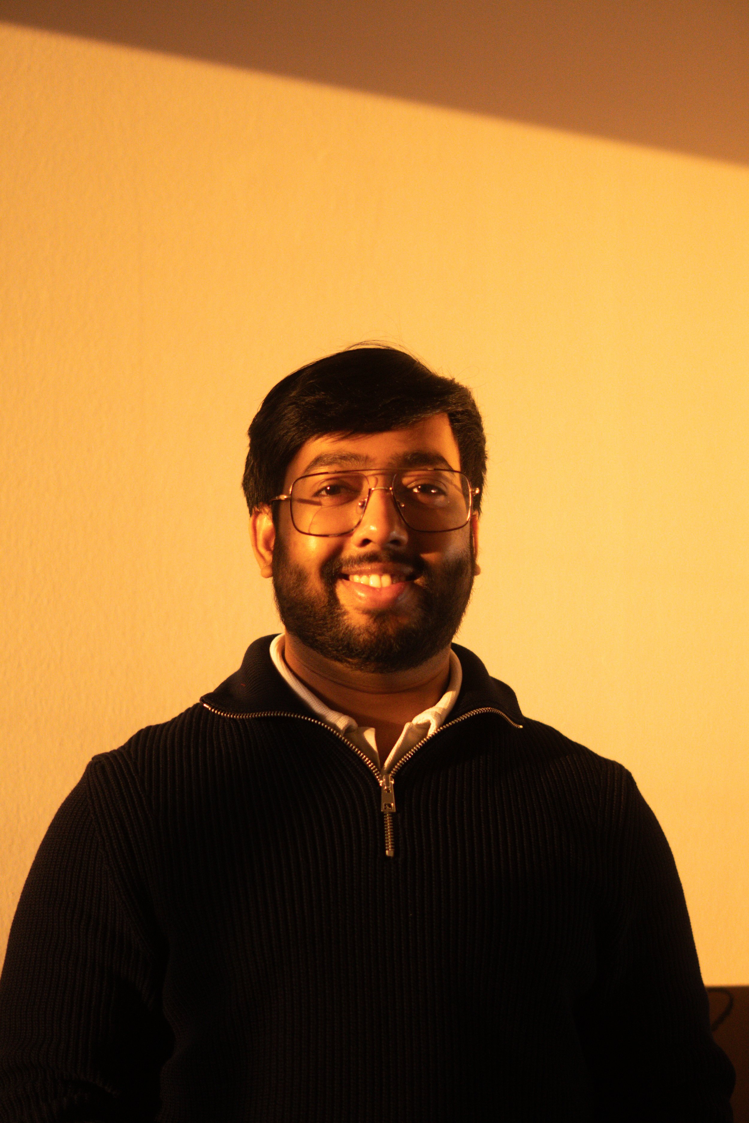 A man with gray hair, wearing a blue sweater and beige pants, sitting on a bench with a neutral expression, casting a shadow on a plain, beige background.