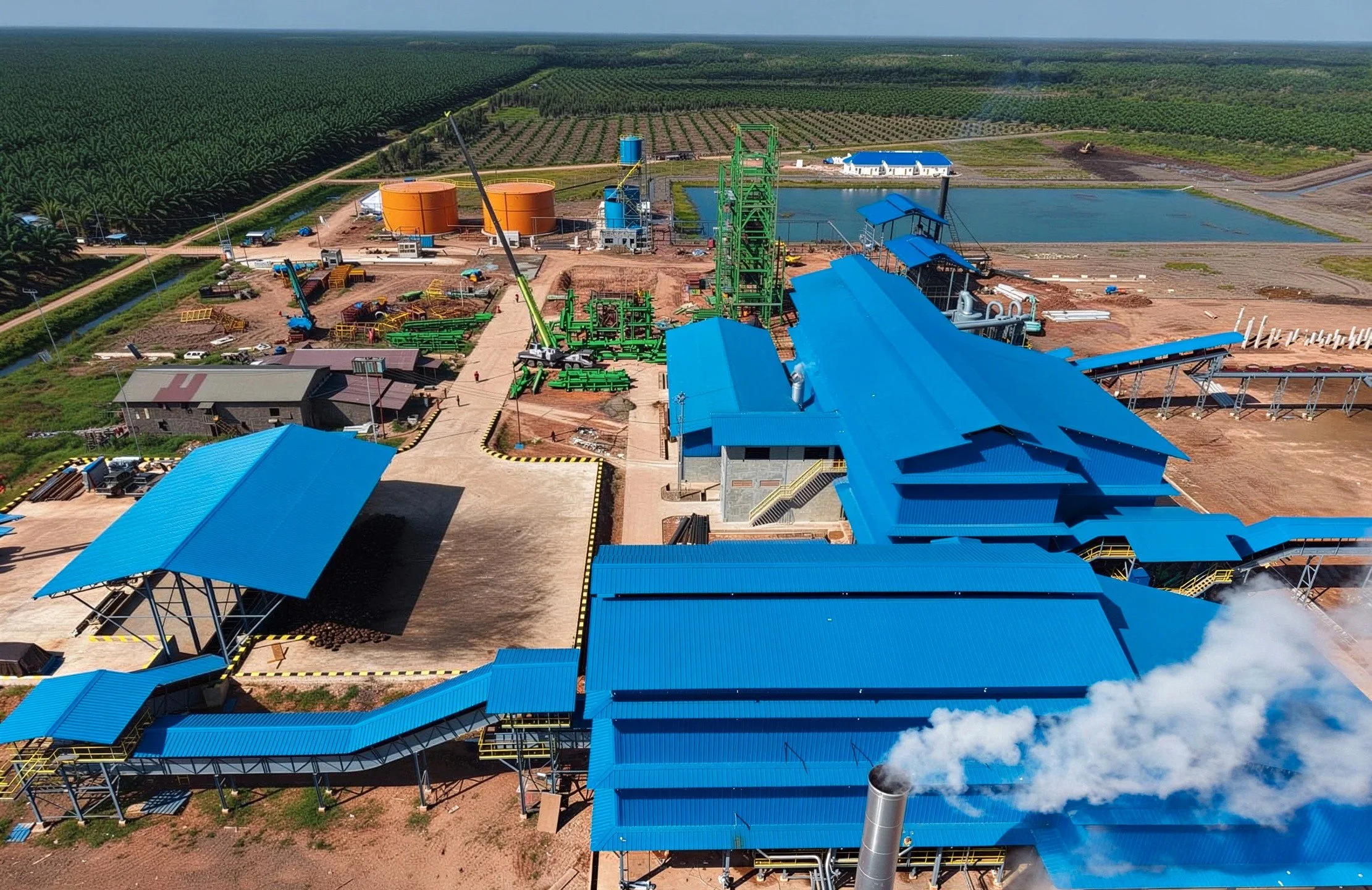 Aerial view of an industrial facility with blue roofs, green structures, tanks, and machinery, adjacent to agricultural fields, with some smoke or steam emitting from a chimney.