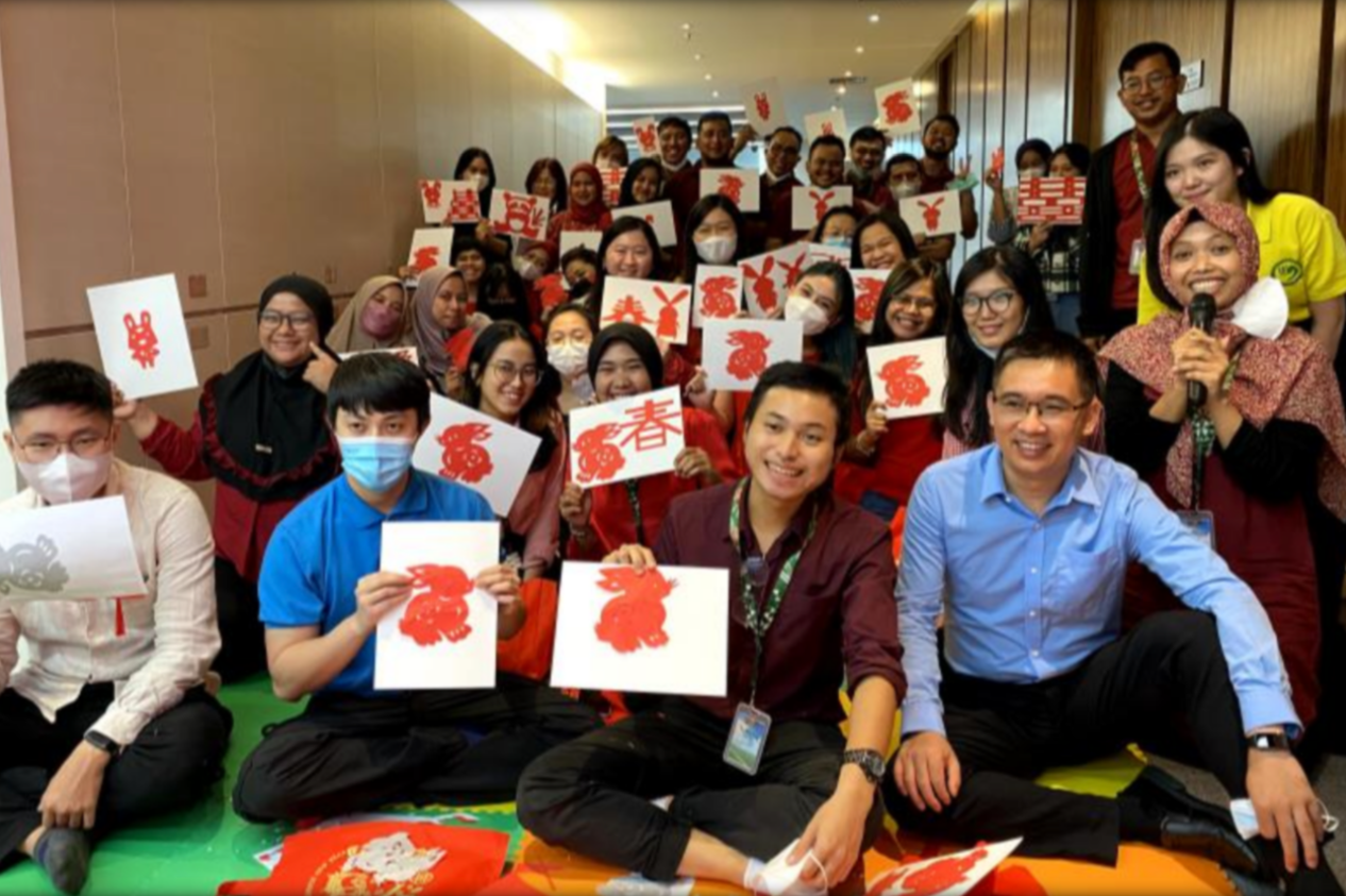 Group of people celebrating Chinese New Year, holding red and white paper cutouts of rabbits and characters, some wearing masks, indoors with beige walls and wooden panels.