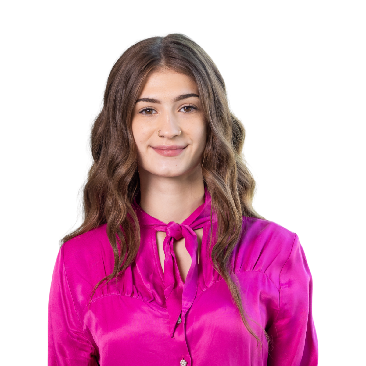 Young woman with brown wavy hair wearing a pink blouse with a bow, standing in front of a white background. 