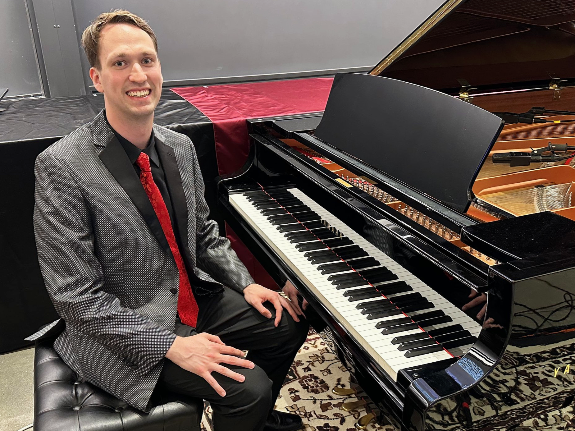 A smiling man seated at a grand piano in an indoor setting, dressed in a patterned blazer, black shirt, and red tie.