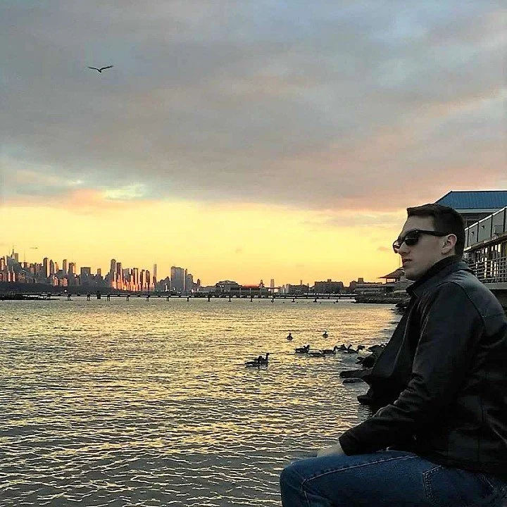 A man wearing sunglasses and a black leather jacket sitting by a river at sunset, with ducks on the water and a city skyline in the background.