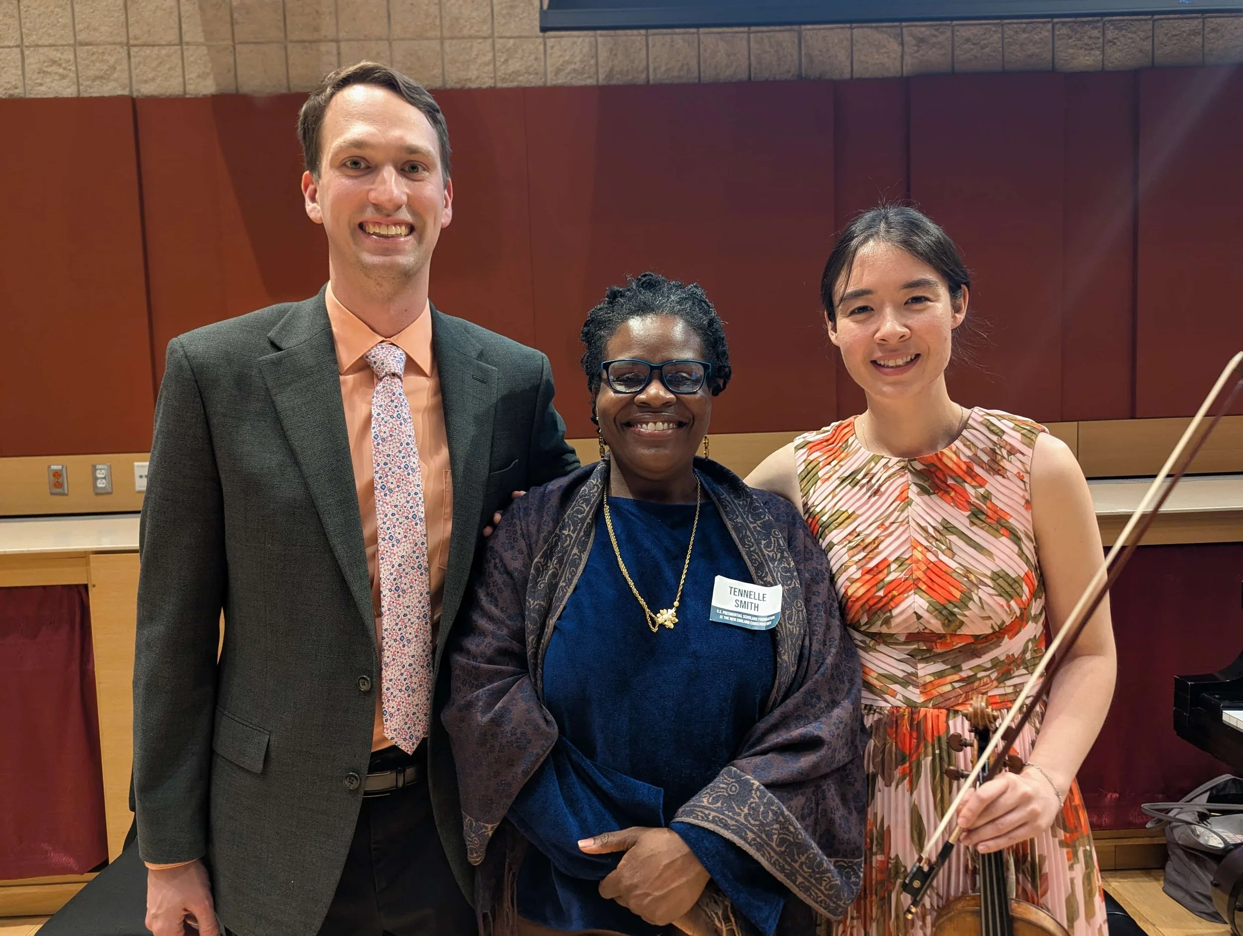 Three women and one man posing together at an indoor event, smiling at the camera. The man is wearing a dark suit with a light pink shirt and patterned tie, the middle woman has glasses, short curly hair, and a blue outfit, and the woman on the right has long dark hair and a floral dress, holding a violin and bow.