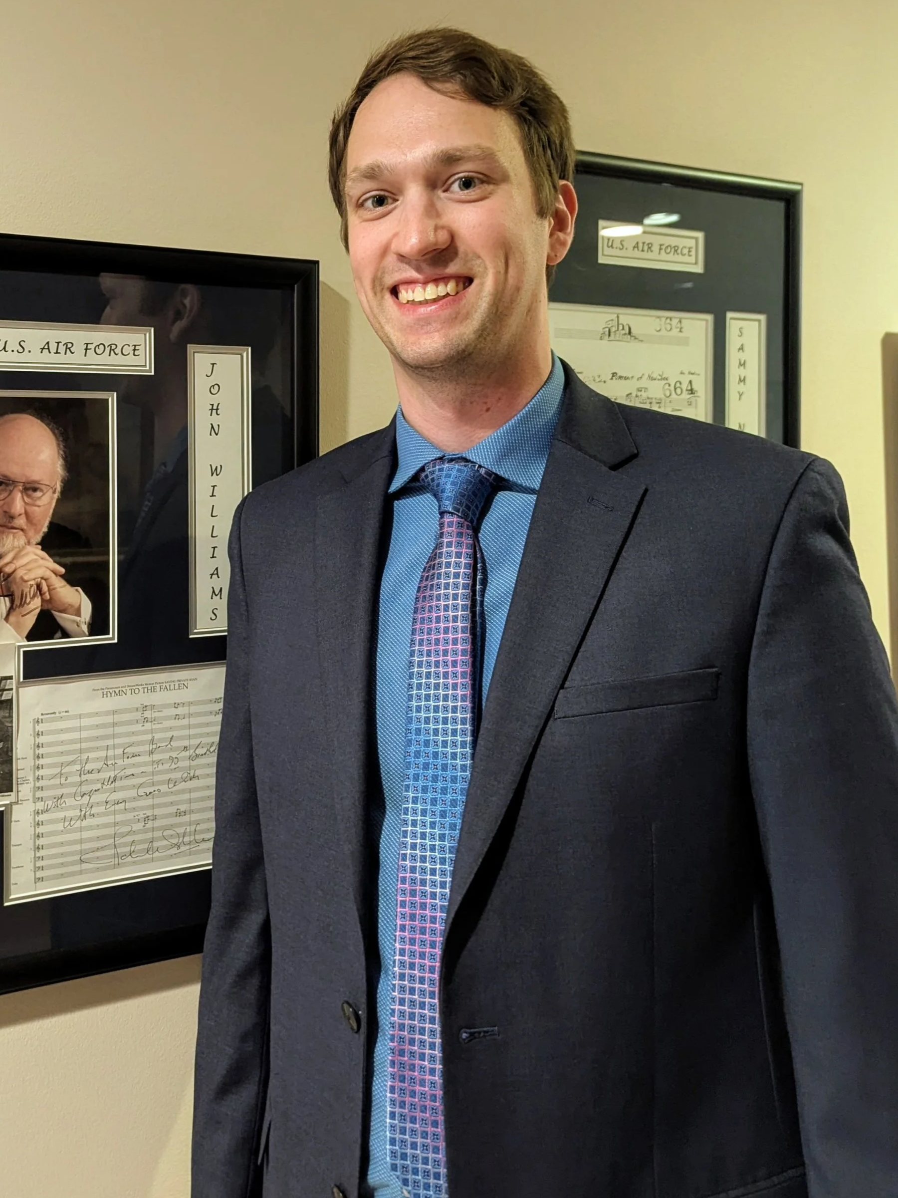 Smiling man in a suit and blue shirt standing near framed documents related to the U.S. Air Force.