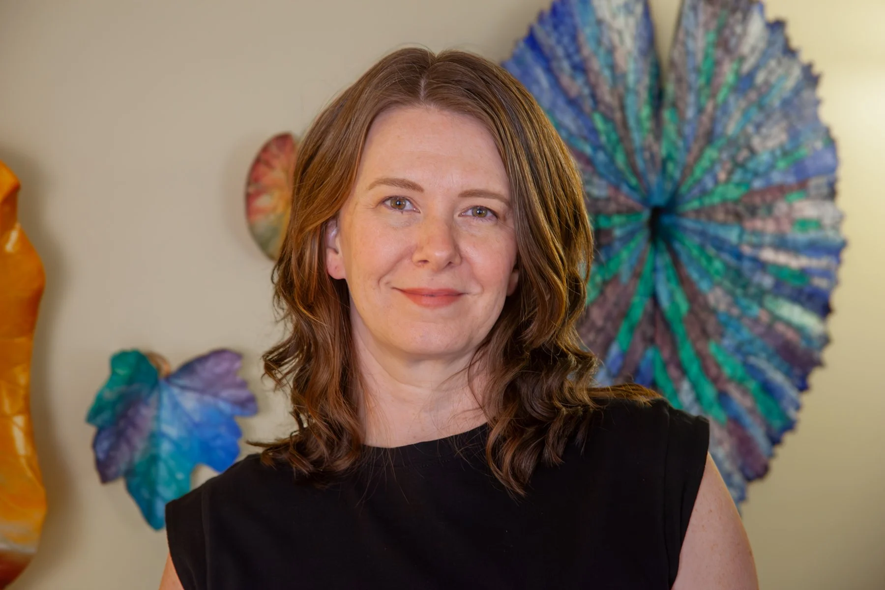 Artist and owner of stone leaf studio smiles at the camera. She has light skin and blue eyes, and is wearing a black top. Behind her are colorful wall art pieces made from large leaf impressions in blue, purple, green, and orange.