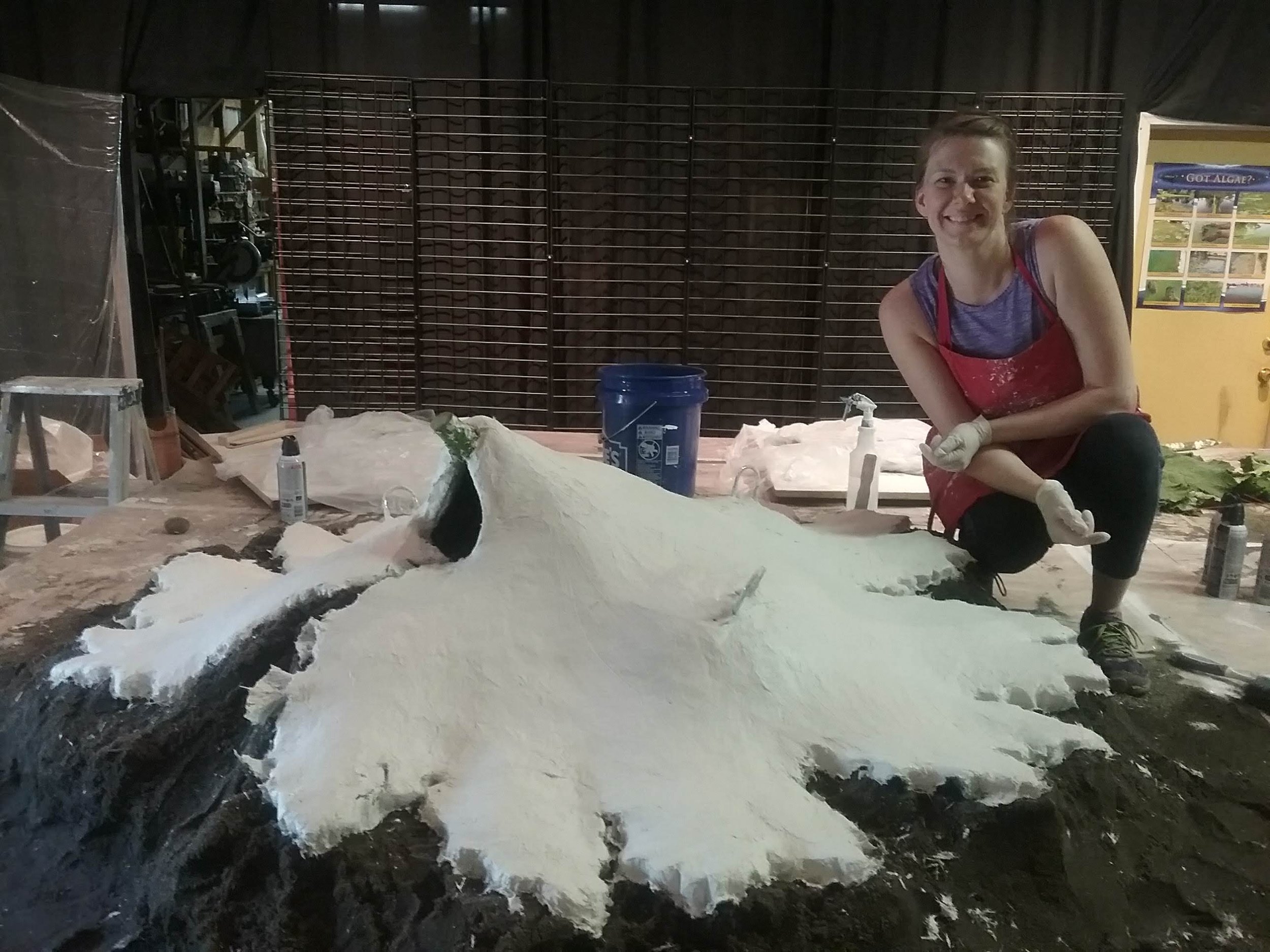 Tania McCamy is crouching next to a large piece of sculpture mold material in a workshop. They are smiling and wearing gloves and an apron.