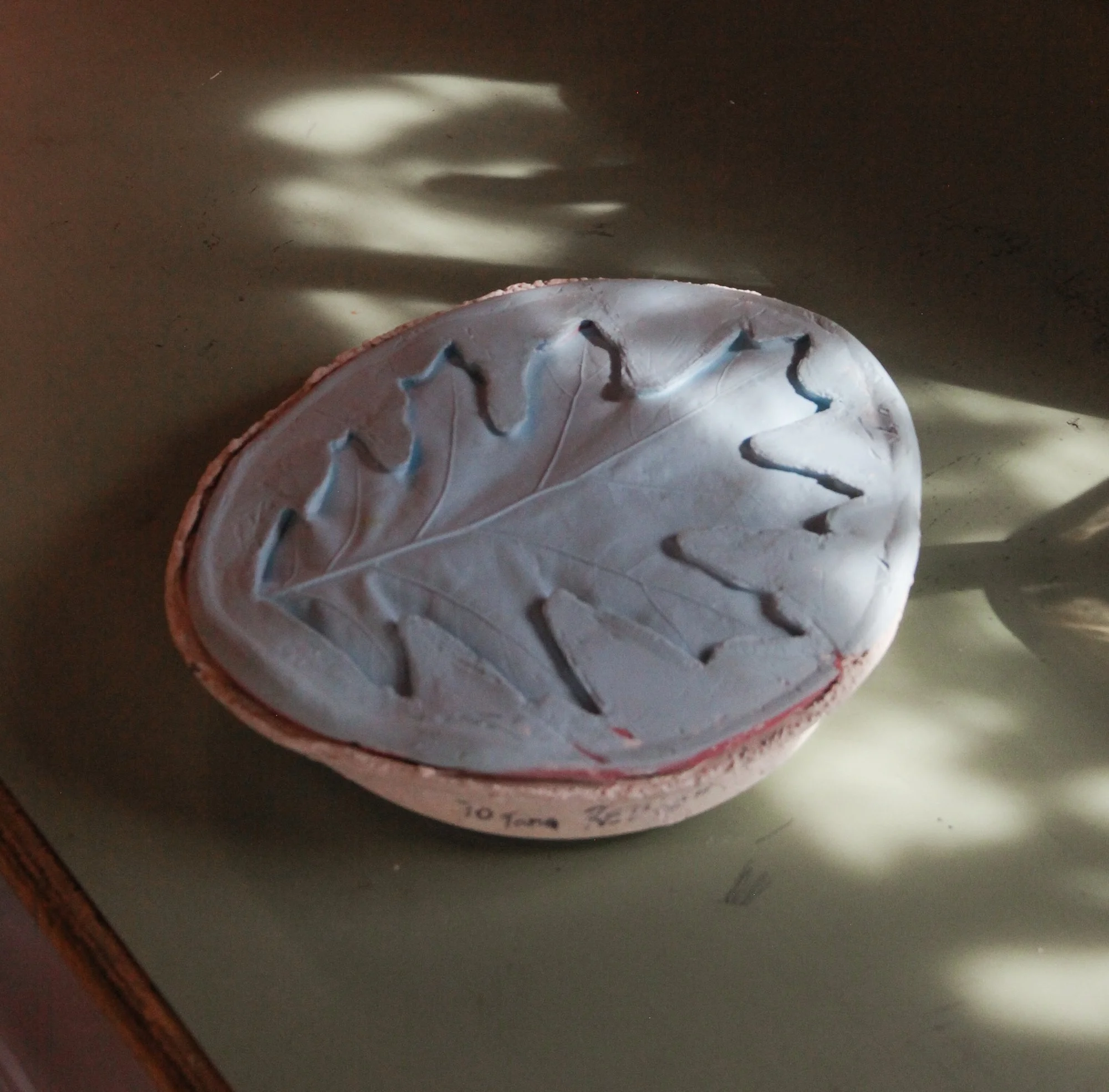 A light blue silicone mold of a leaf sits on a sage green tabletop that is dappled with sunlight.