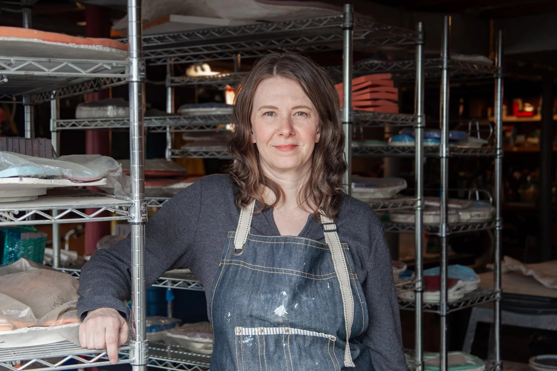 Artist Tania McCamy with shoulder-length brown hair, wearing a dark shirt and a denim apron, stands in her art studio infront of metal shelves filled with plates, trays, and supplies in the background.