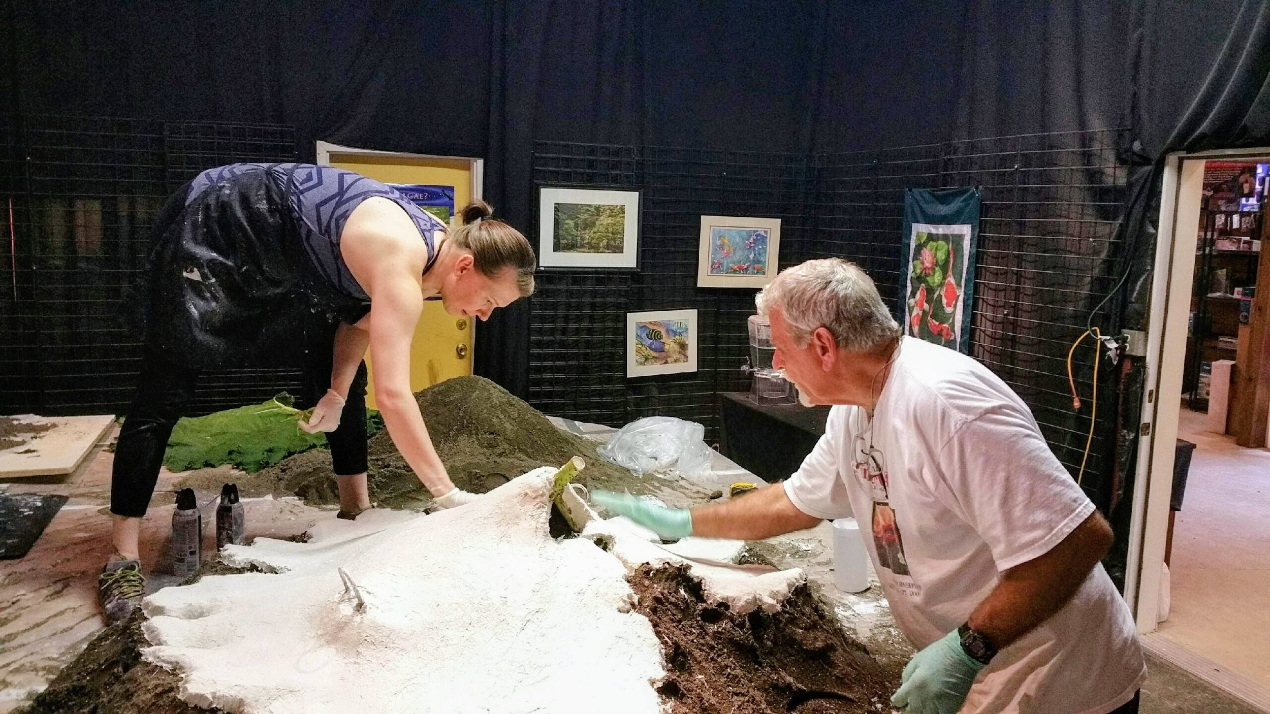 Two women and a man working on an art project that involves a sandy or soil-like material on a large flat surface in a room with black walls and framed artwork.